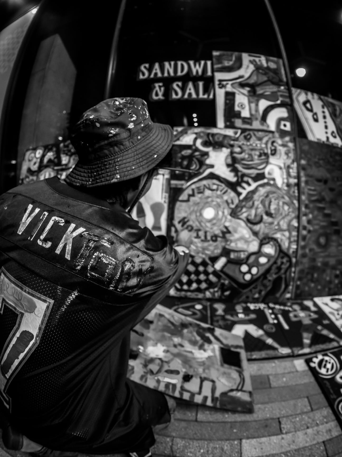 Person wearing a hat and jacket with 'VICK' on the back, looking at colorful artwork outside a sandwich shop.
