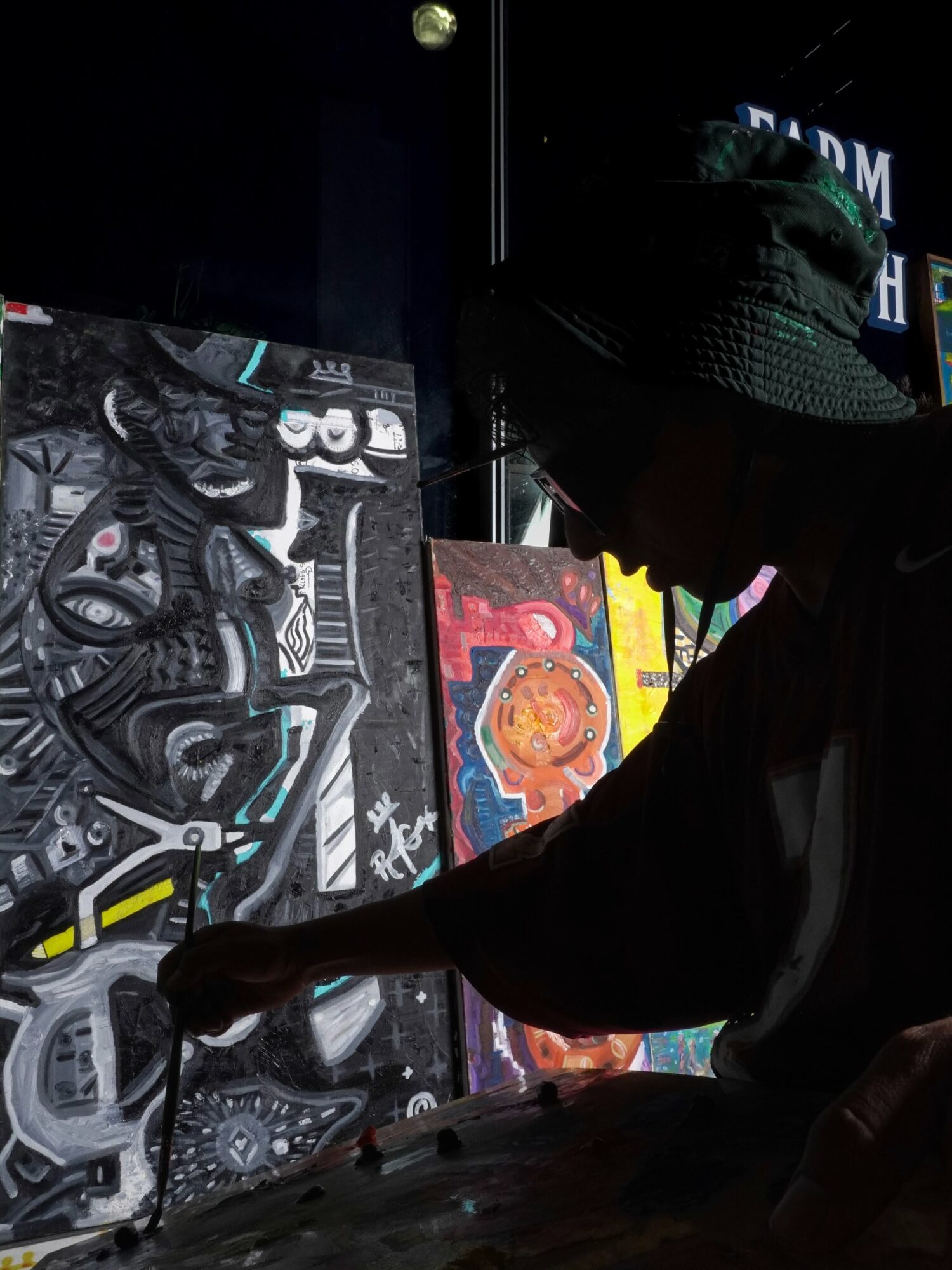 Person wearing a hat and dark clothing looking at colorful artwork on display, with a dark background.