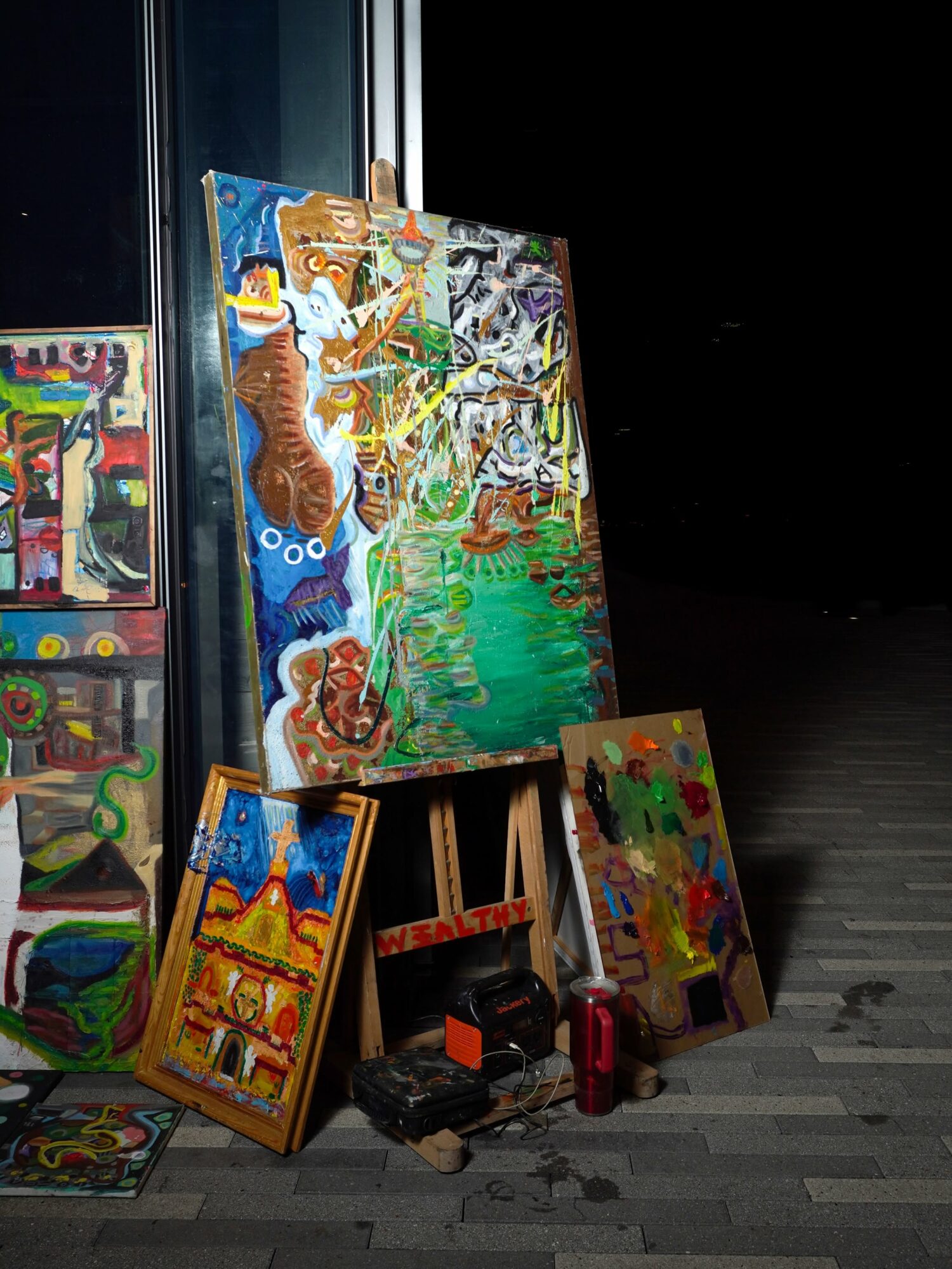 Paintings leaning against a wall, with a black background, some art supplies, and a red water bottle on the floor.