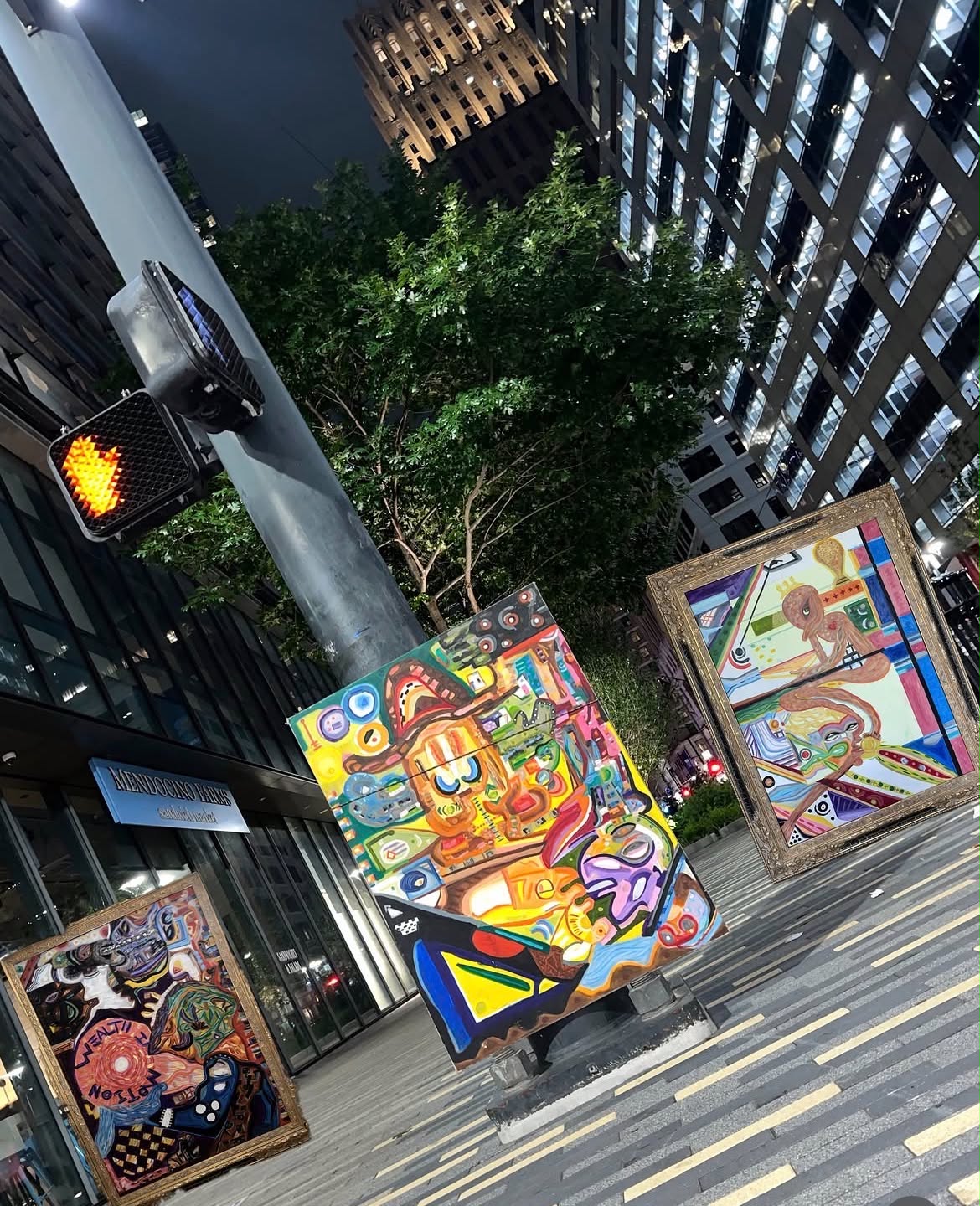 Street scene with colorful abstract art displays, tall buildings, a traffic light, and a tree in an urban setting.