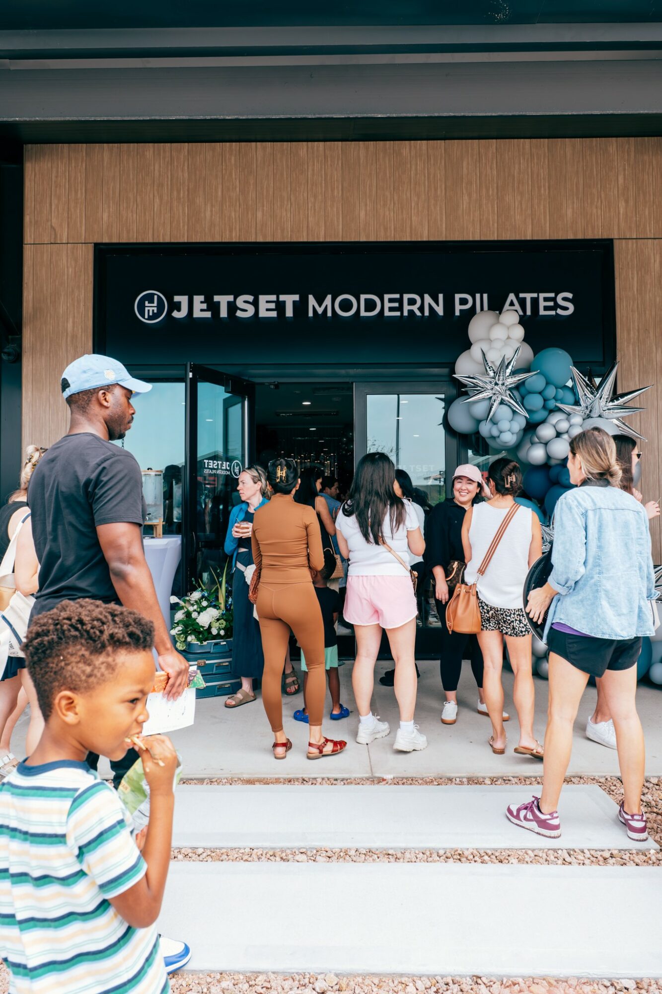 People entering Jetset Modern Pilates studio, some wearing masks, children and adults, outside storefront with balloons, wooden facade.