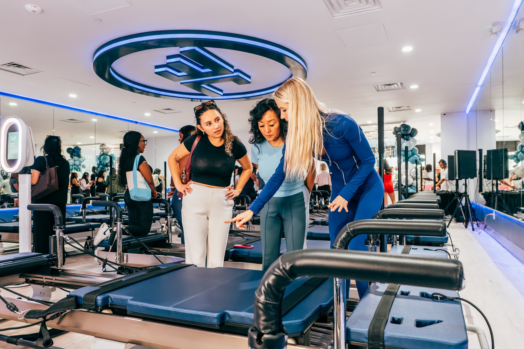 Two women in workout clothes look at a treadmill in a brightly lit gym with mirrors and exercise equipment.