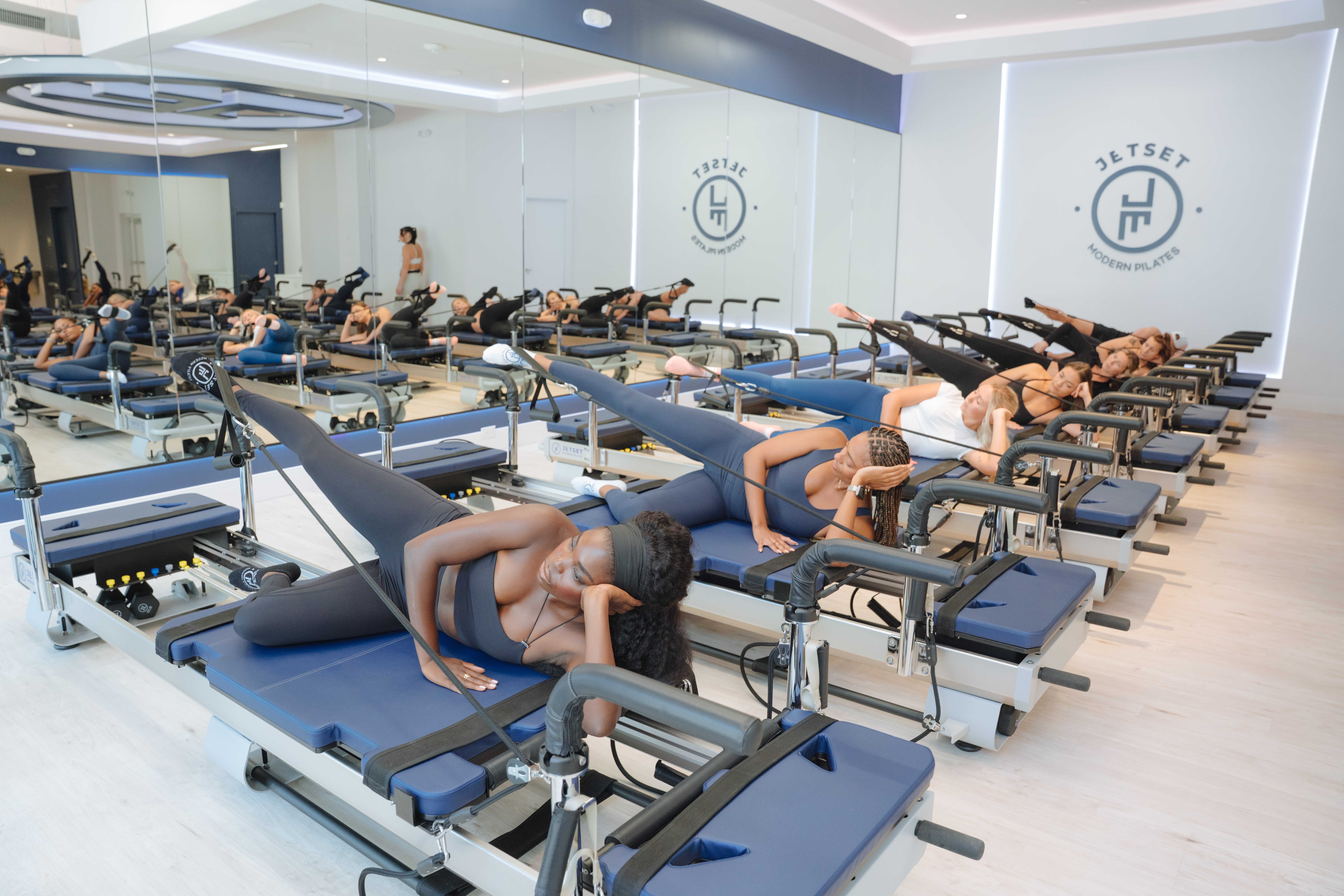 Group of people exercising on Pilates reformer machines in a fitness studio with white walls and logos.