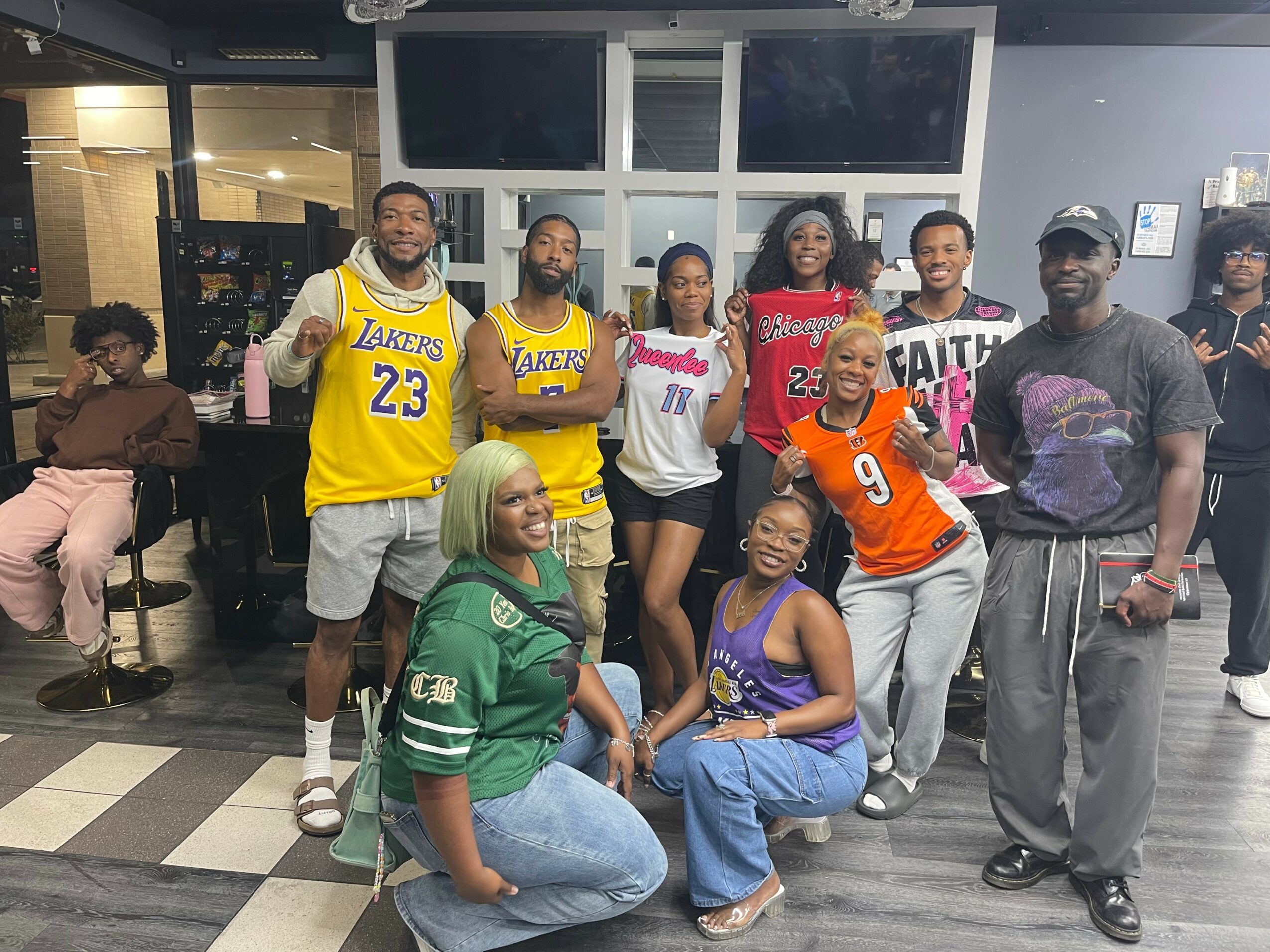 Group of nine people, some wearing sports jerseys, standing and sitting indoors with large windows behind them.