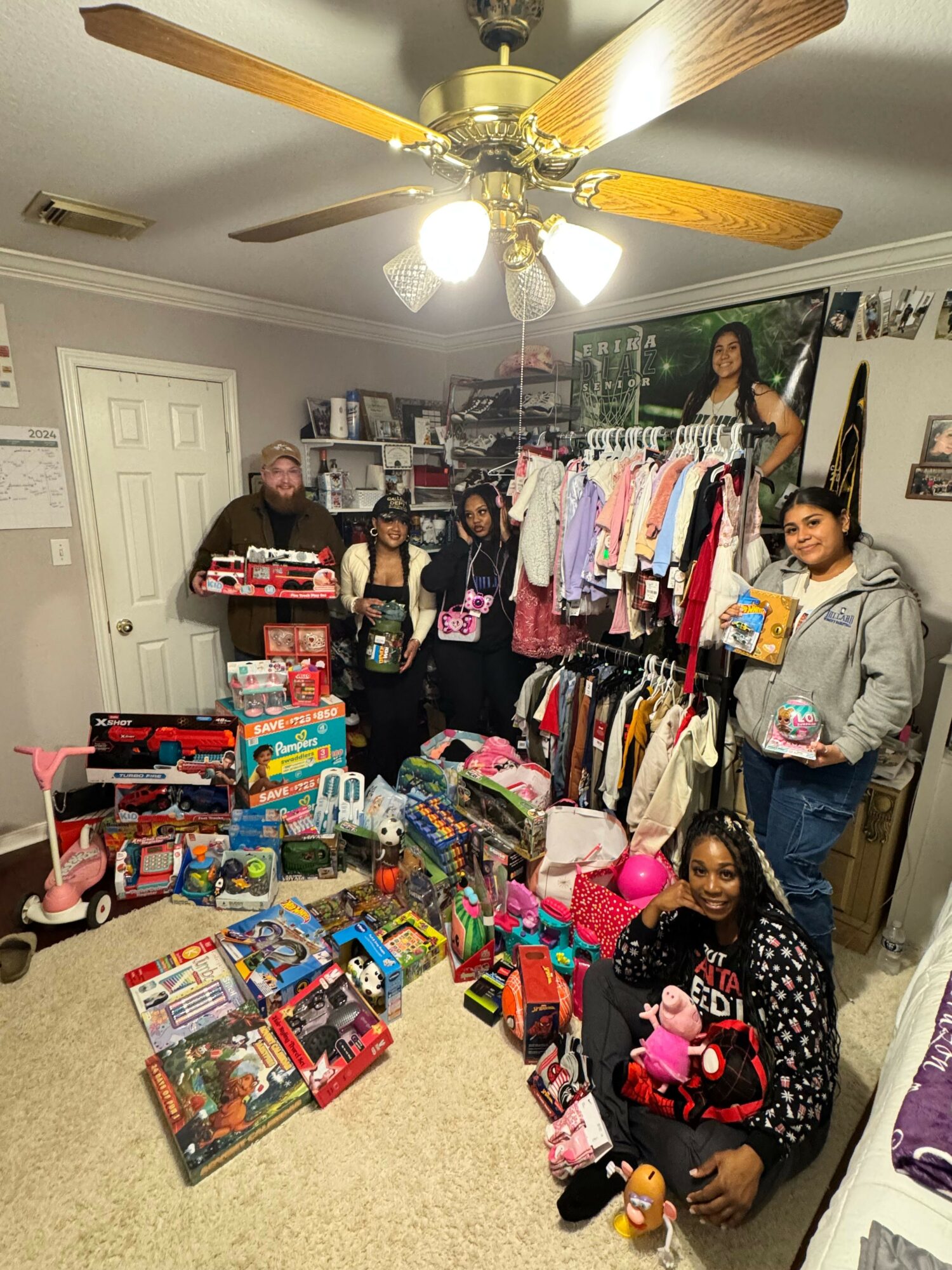 Group of people in a room with shelves and toys, some holding gifts, with a ceiling fan overhead.
