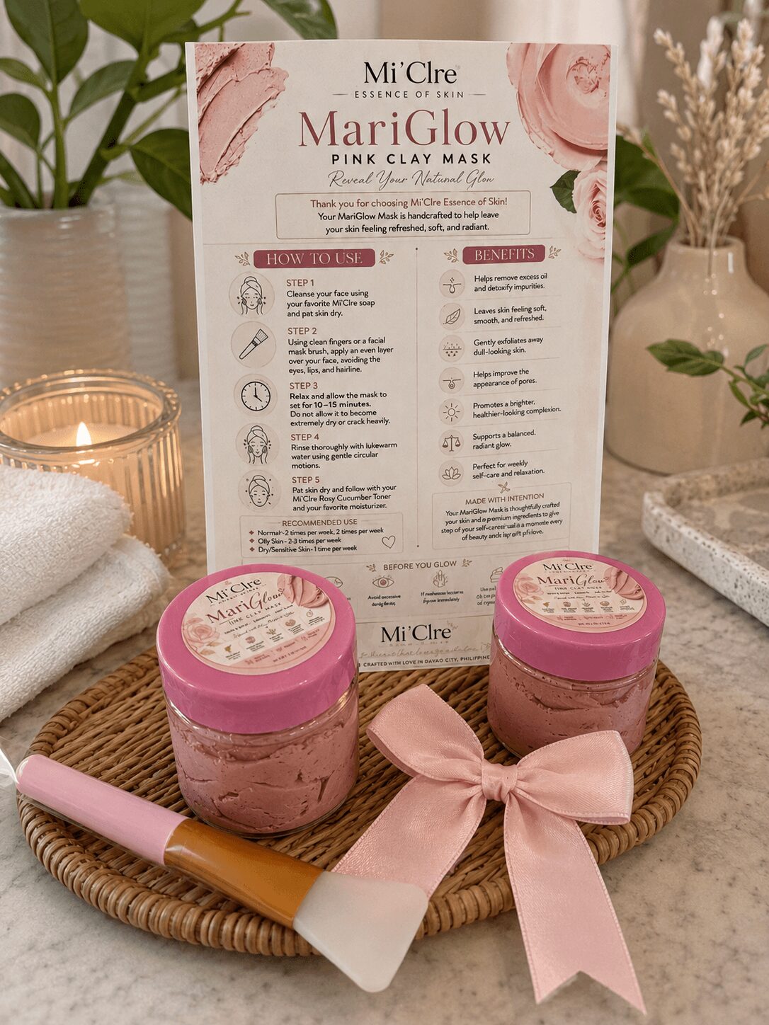 Two pink jars of clay masks on a tray with a pink ribbon, a brush, and a skincare instruction sheet, surrounded by candles and plants.
