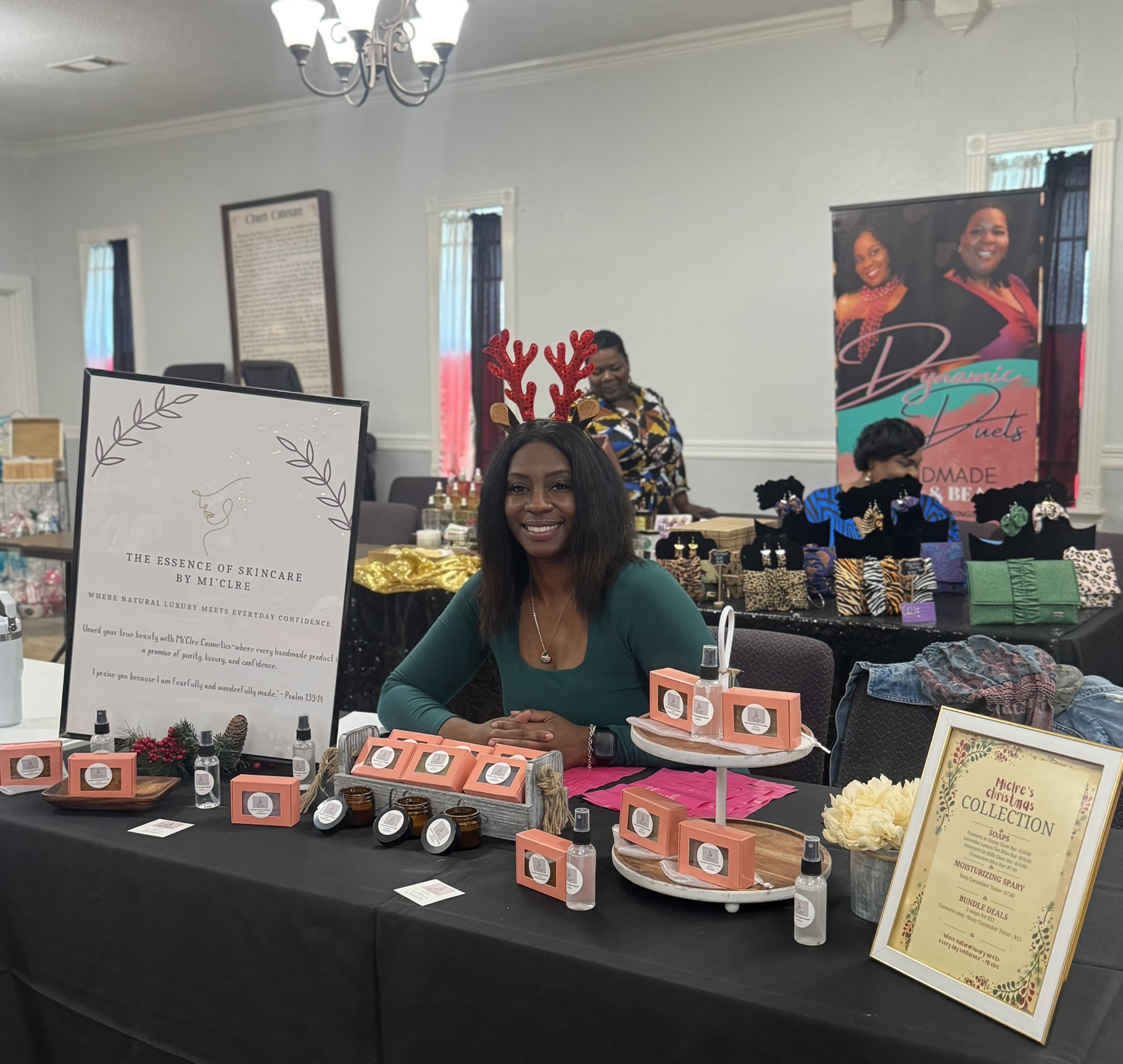 Smiling woman wearing reindeer antlers headband sits at a table with jewelry displays, in a room with promotional posters and people.