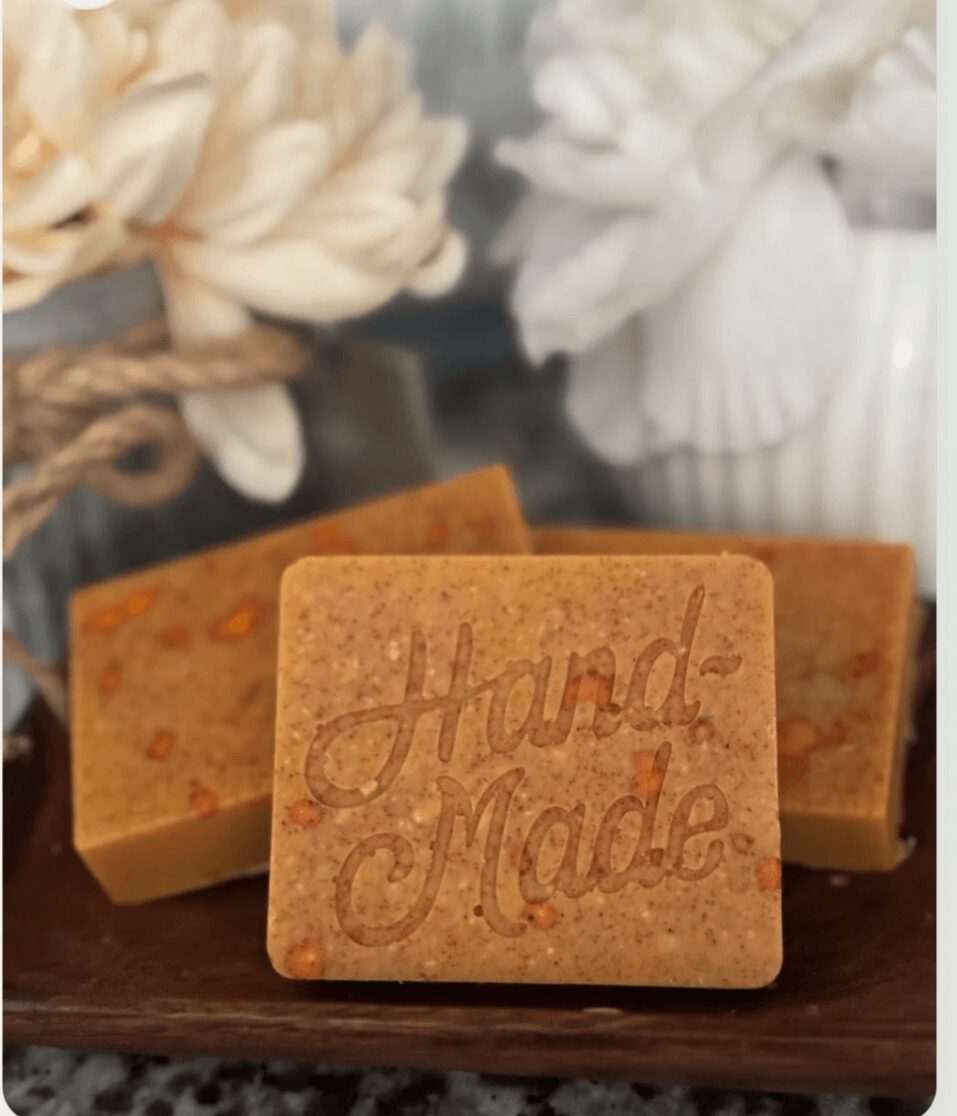 Three handmade soap bars on a wooden surface with flowers and a white vase in the background.