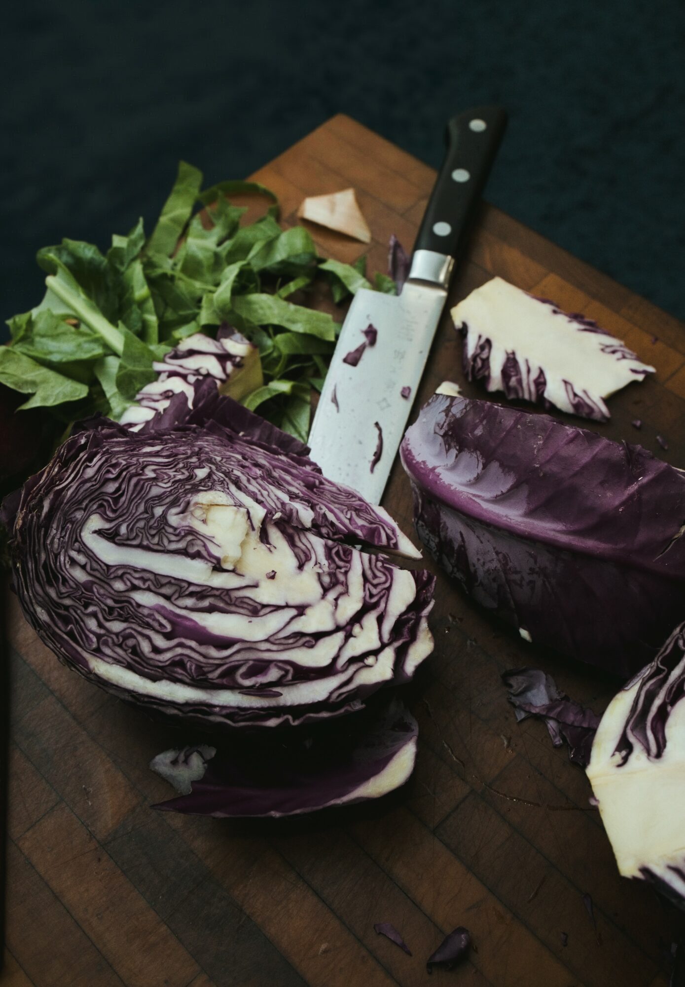 Half-cut purple cabbage with green leafy vegetables and a knife on a wooden cutting board.