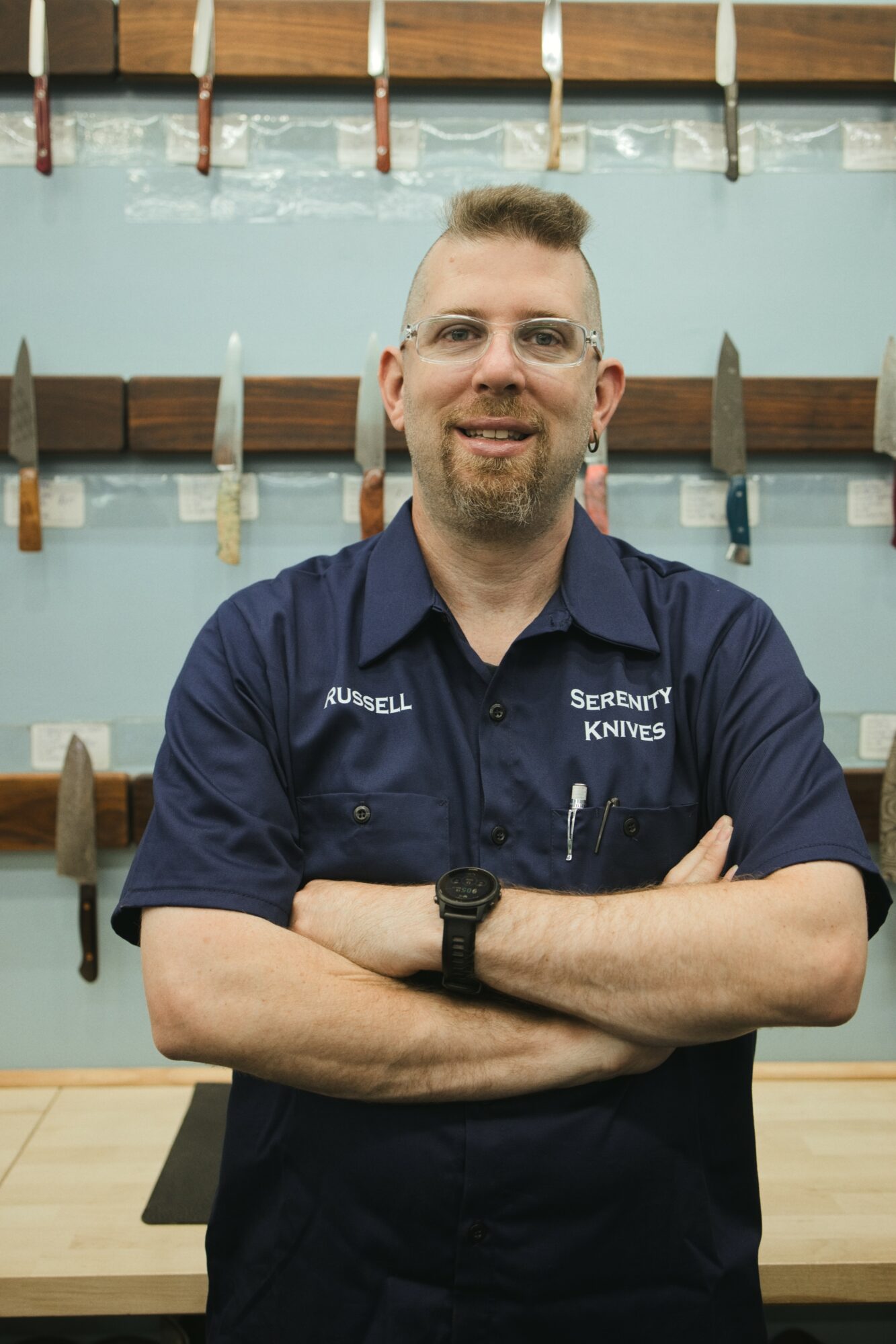 Man with glasses and a beard, wearing a navy shirt, standing with arms crossed in front of a display of knives on a wall.