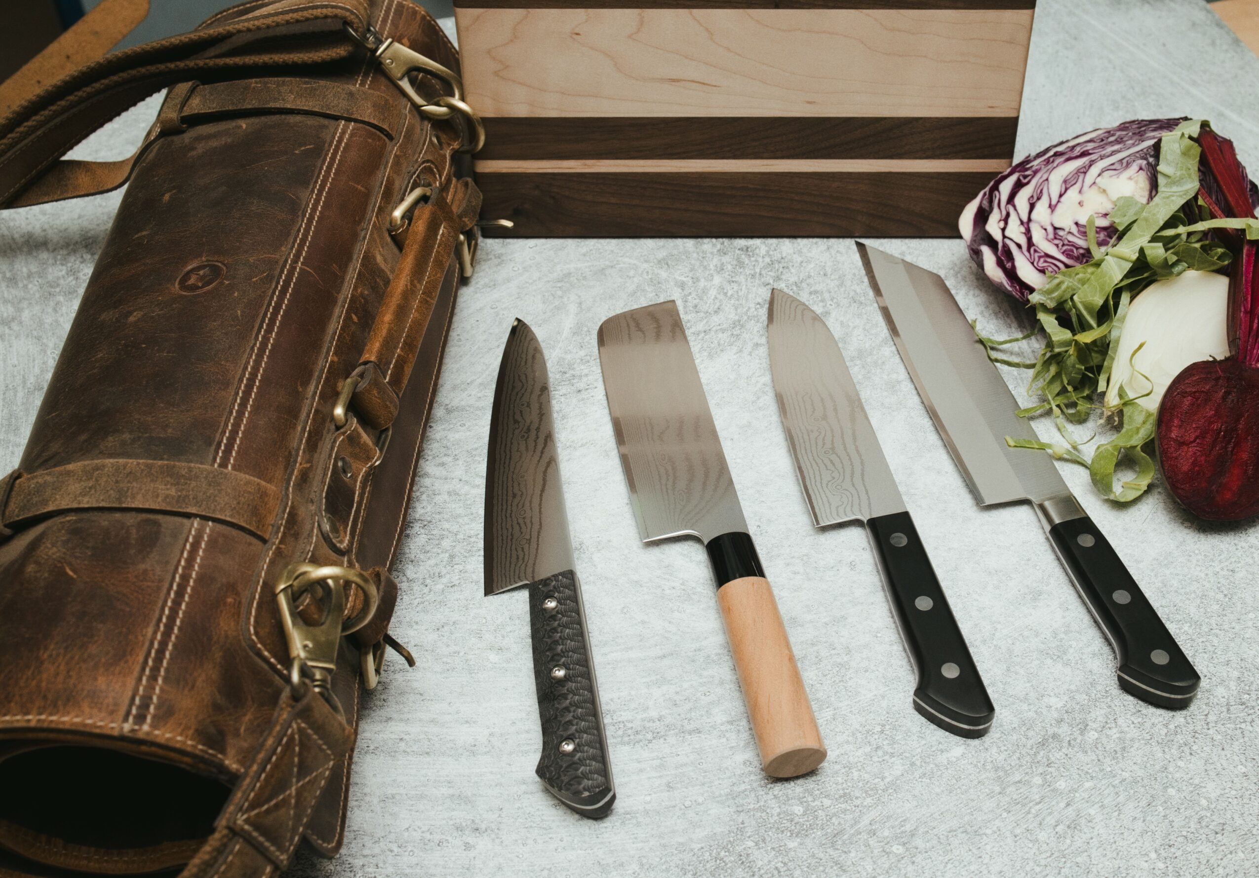 Five knives with black handles and a wooden handle, a bouquet of flowers, and a leather bag on a light surface.