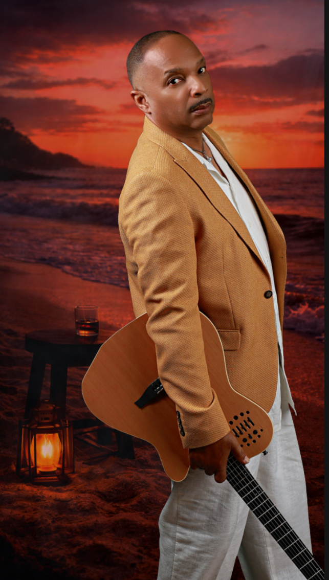 Person holding a guitar on a beach at sunset, wearing a tan blazer and white shirt.