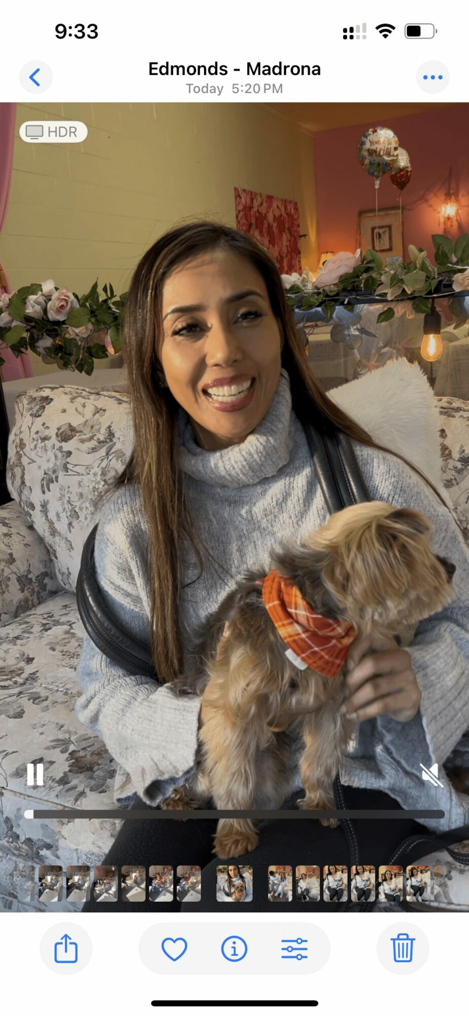 Woman smiling holding a small dog in a decorated room with flowers and artwork behind her.