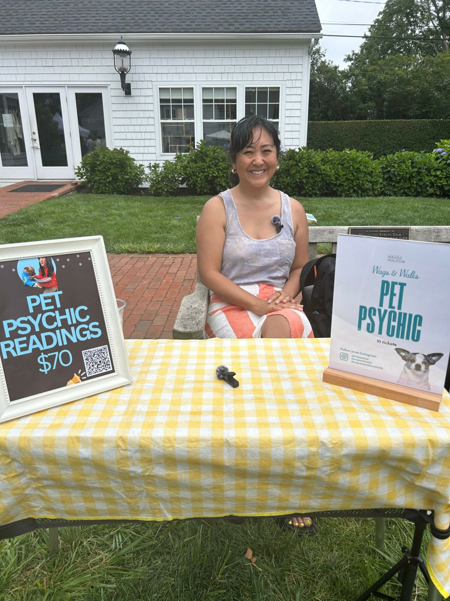 A woman stands outdoors near a table with books and a yellow checkered tablecloth, holding a book titled 'The Psyc of Psychedelic' with a dog illustration. The scene is in a garden with a white house in the background.
