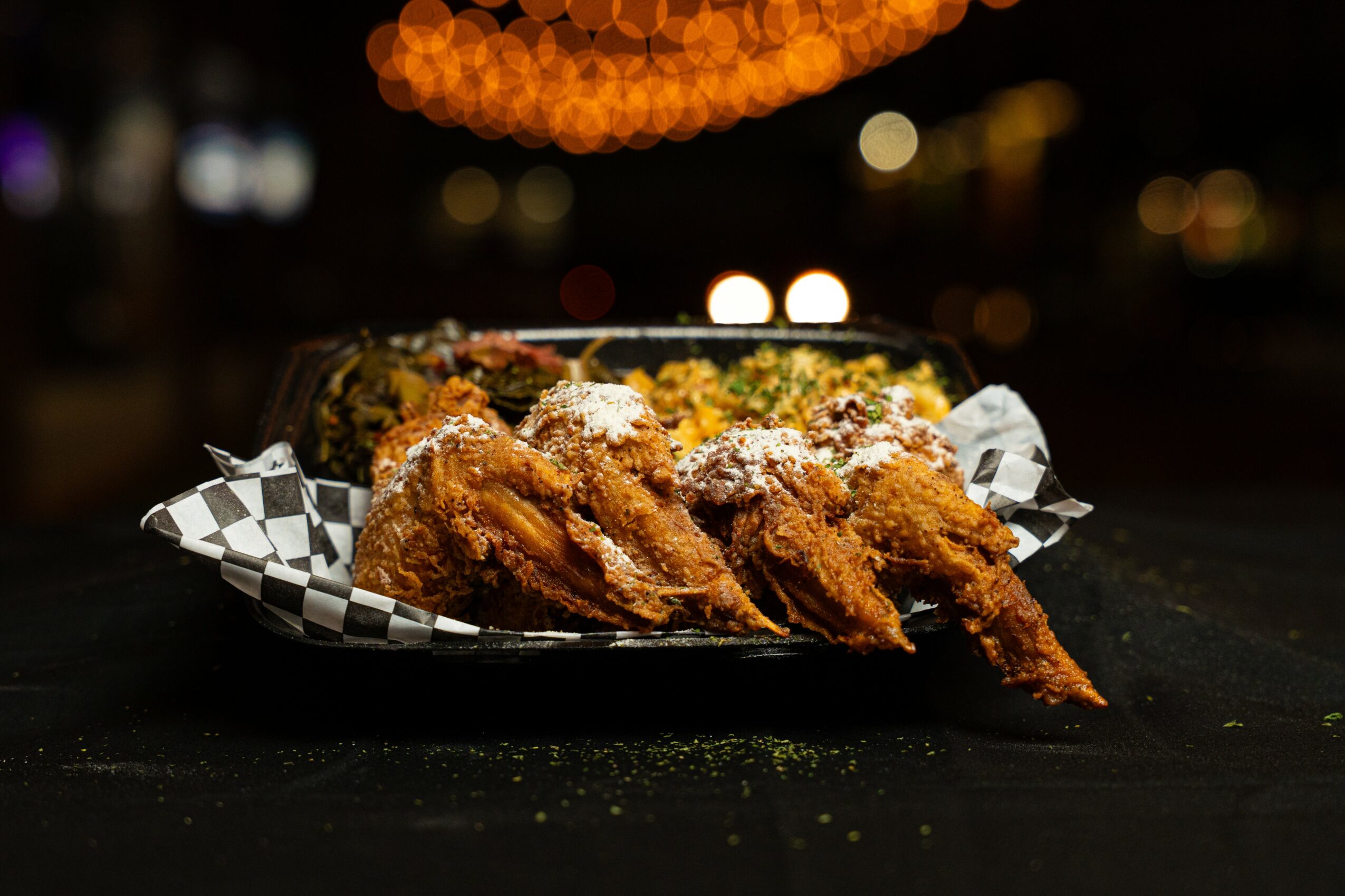 Fried chicken pieces on checkered paper in a black container, with a blurred night city background.