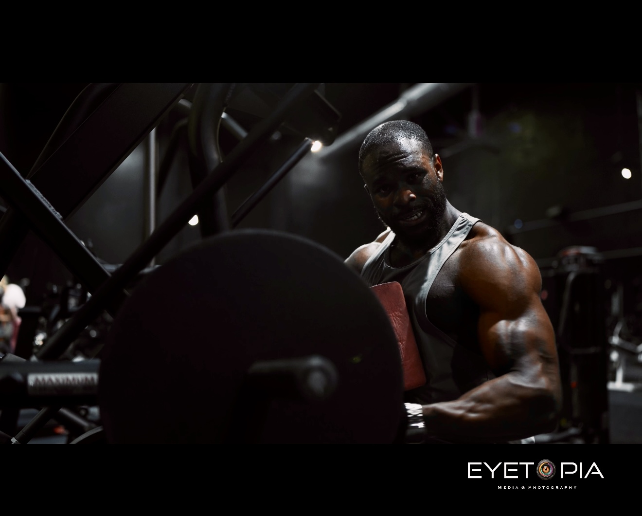 A muscular man lifting weights in a gym, with dark lighting and equipment in the background.