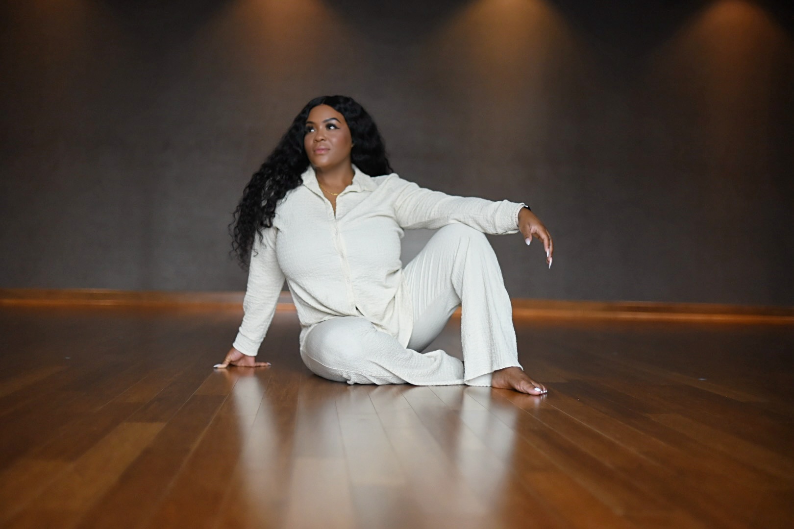 Woman in white outfit sitting on wooden floor with dark background, looking to the side.