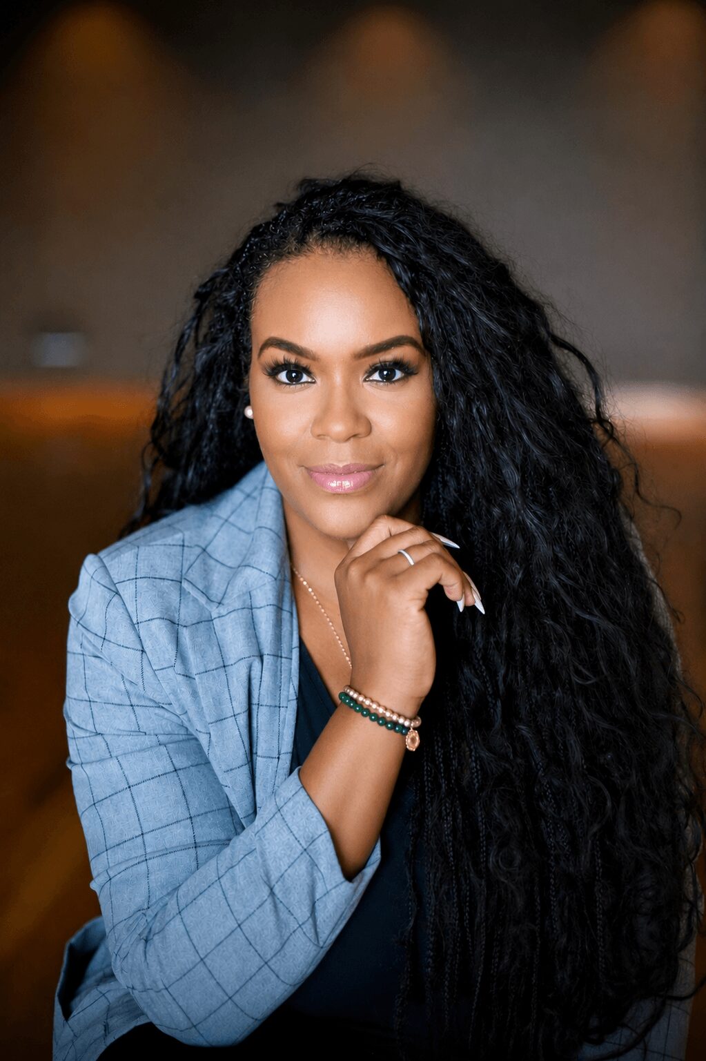 Woman with long curly hair wearing a blue blazer, sitting indoors with a blurred background.