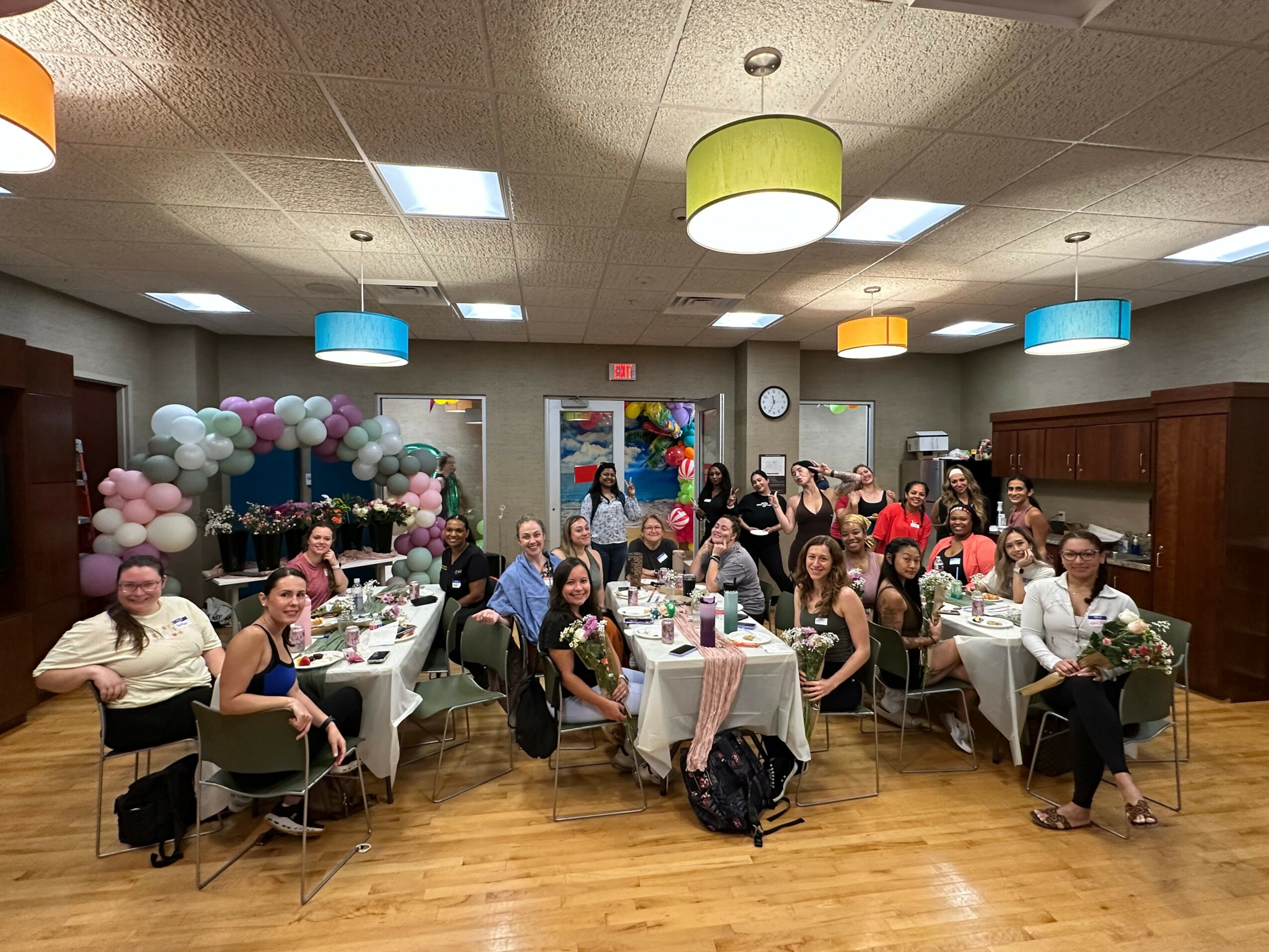 Group of people seated at tables in a decorated room with balloons and colorful hanging lights.