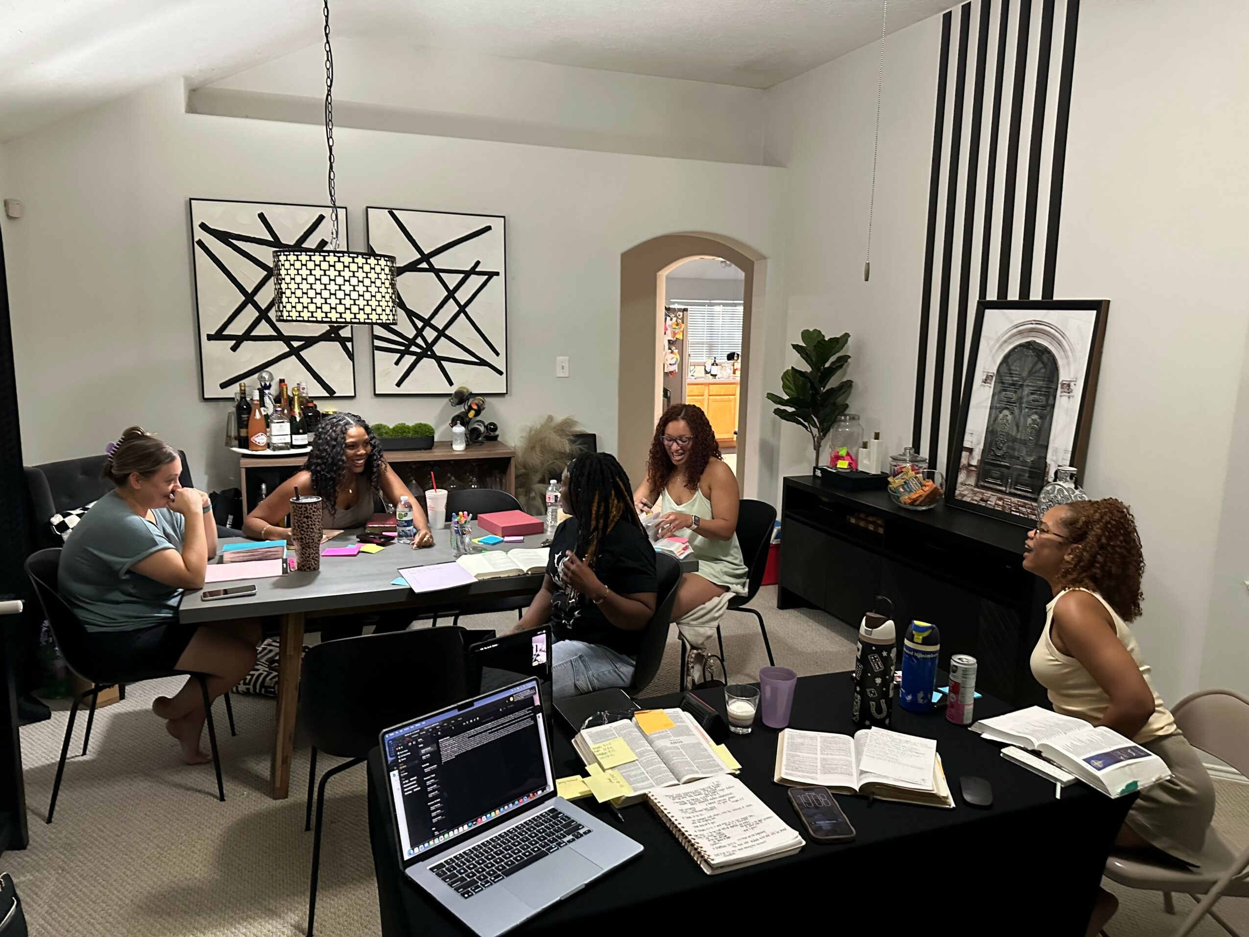 Six people sit around a table in a room with white walls, books, and a laptop, engaged in conversation or reading.