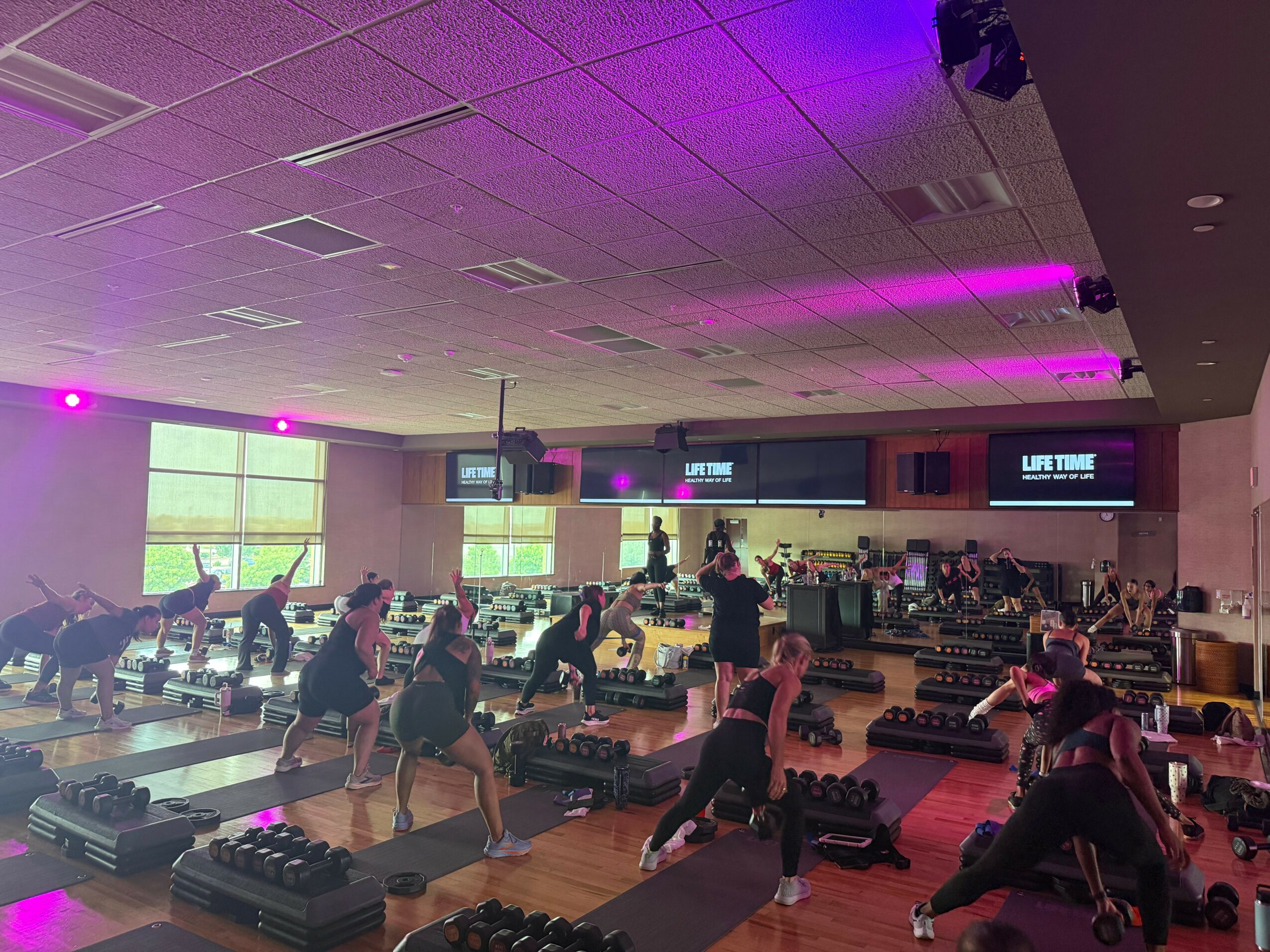 People participating in a group fitness class in a gym, lifting weights and stretching, with screens and windows in the background.