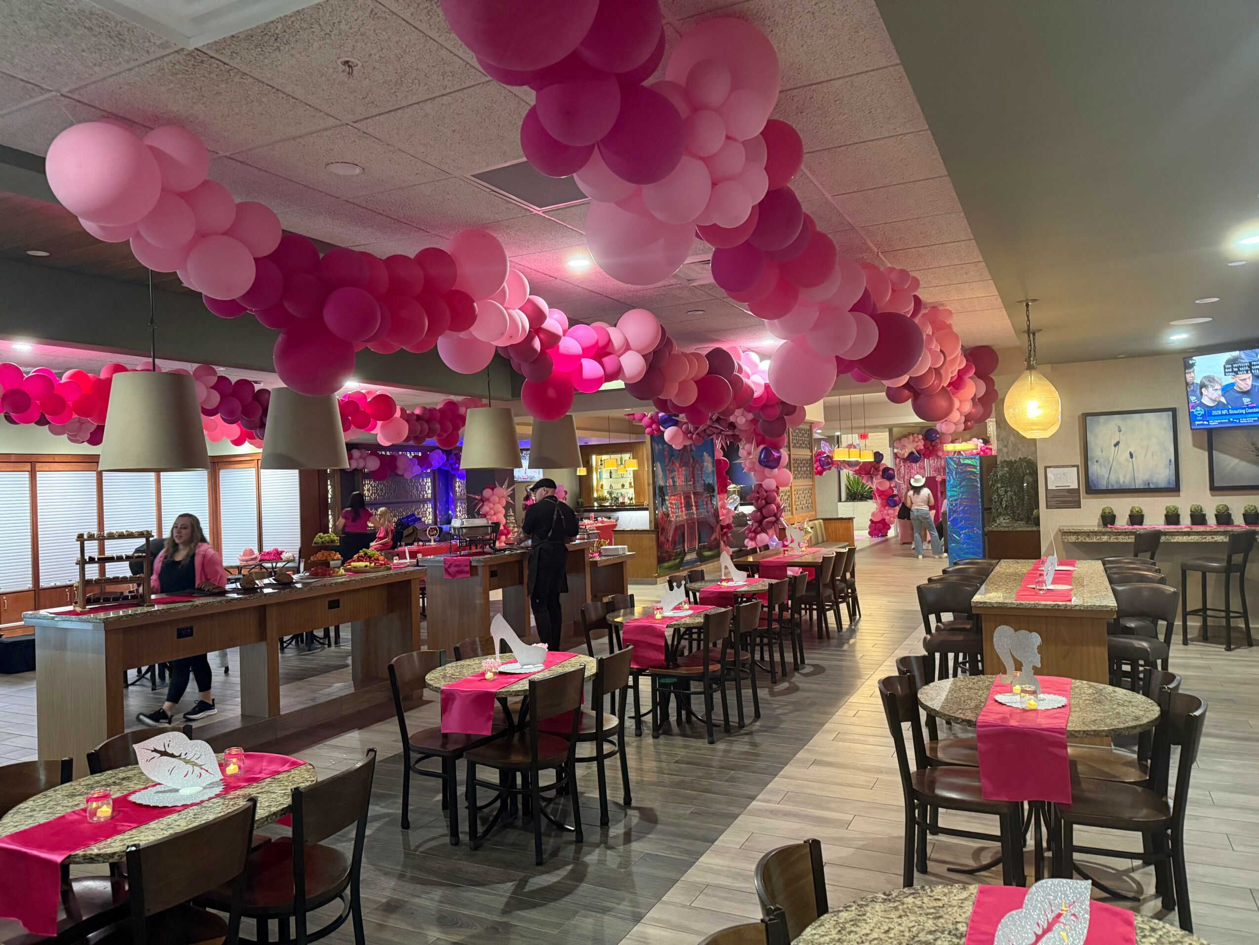 Restaurant decorated with pink balloons hanging from ceiling, tables with pink tablecloths, and chairs arranged around tables.