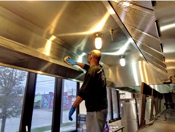 Person cleaning a large stainless steel kitchen hood with a cloth, inside a commercial kitchen with windows showing outdoor scenery.