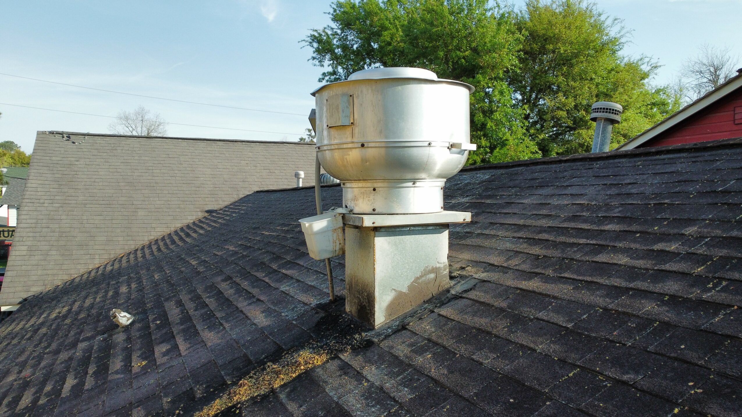Metal chimney pipe on a sloped roof with shingles, surrounded by trees and sky.