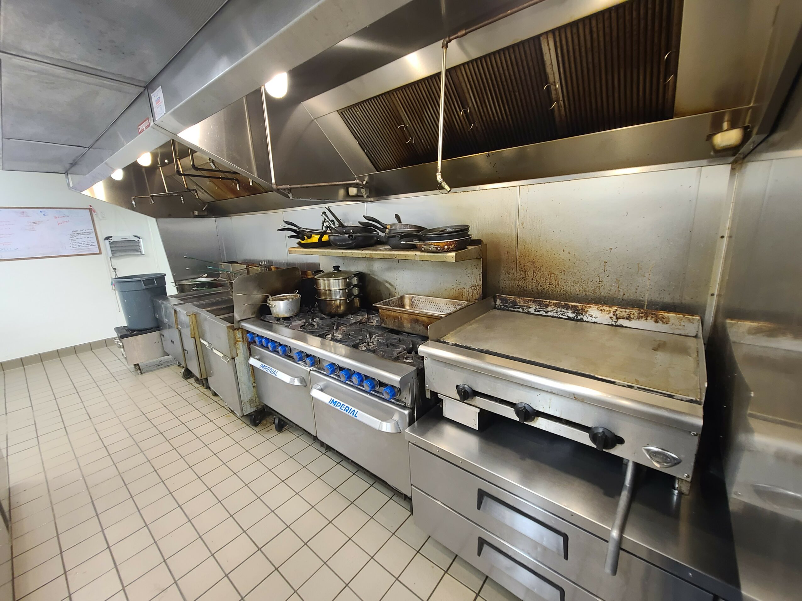 Commercial kitchen with stove, grill, and stainless steel surfaces, tiled floor, and overhead ventilation system.