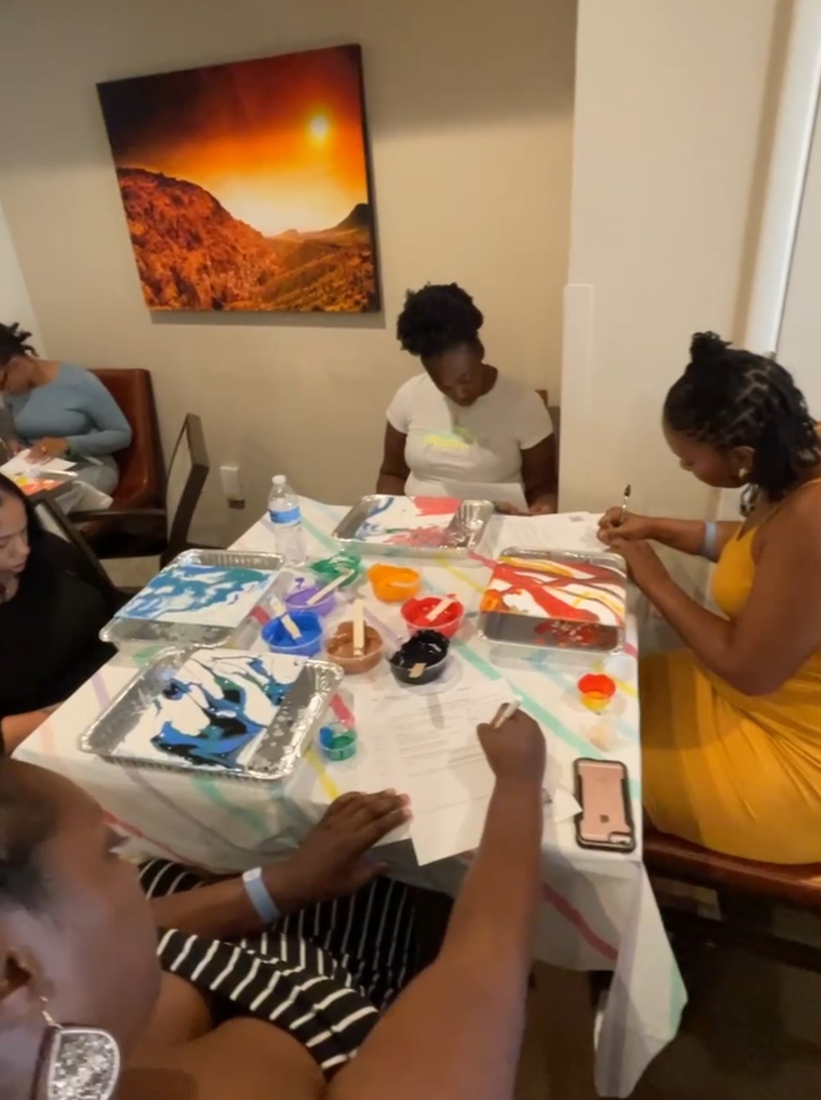 People sitting around a table with art supplies, painting and drawing, in a room with a landscape painting on the wall.