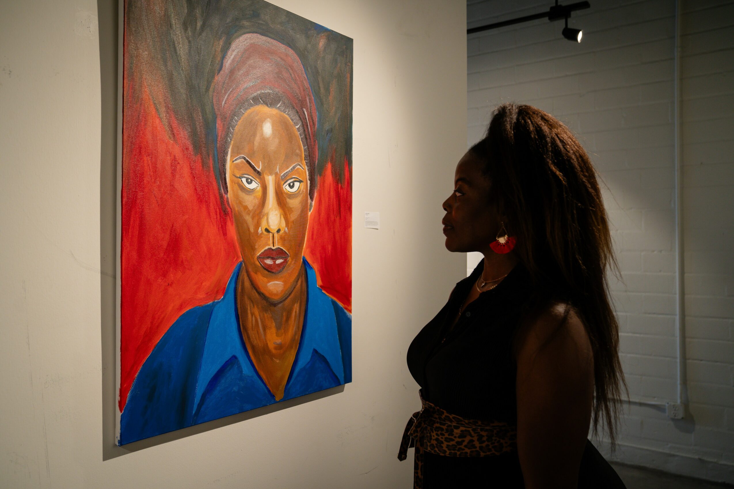A woman with long hair looks at a portrait of a person with a headscarf, painted with bold colors and expressive style.