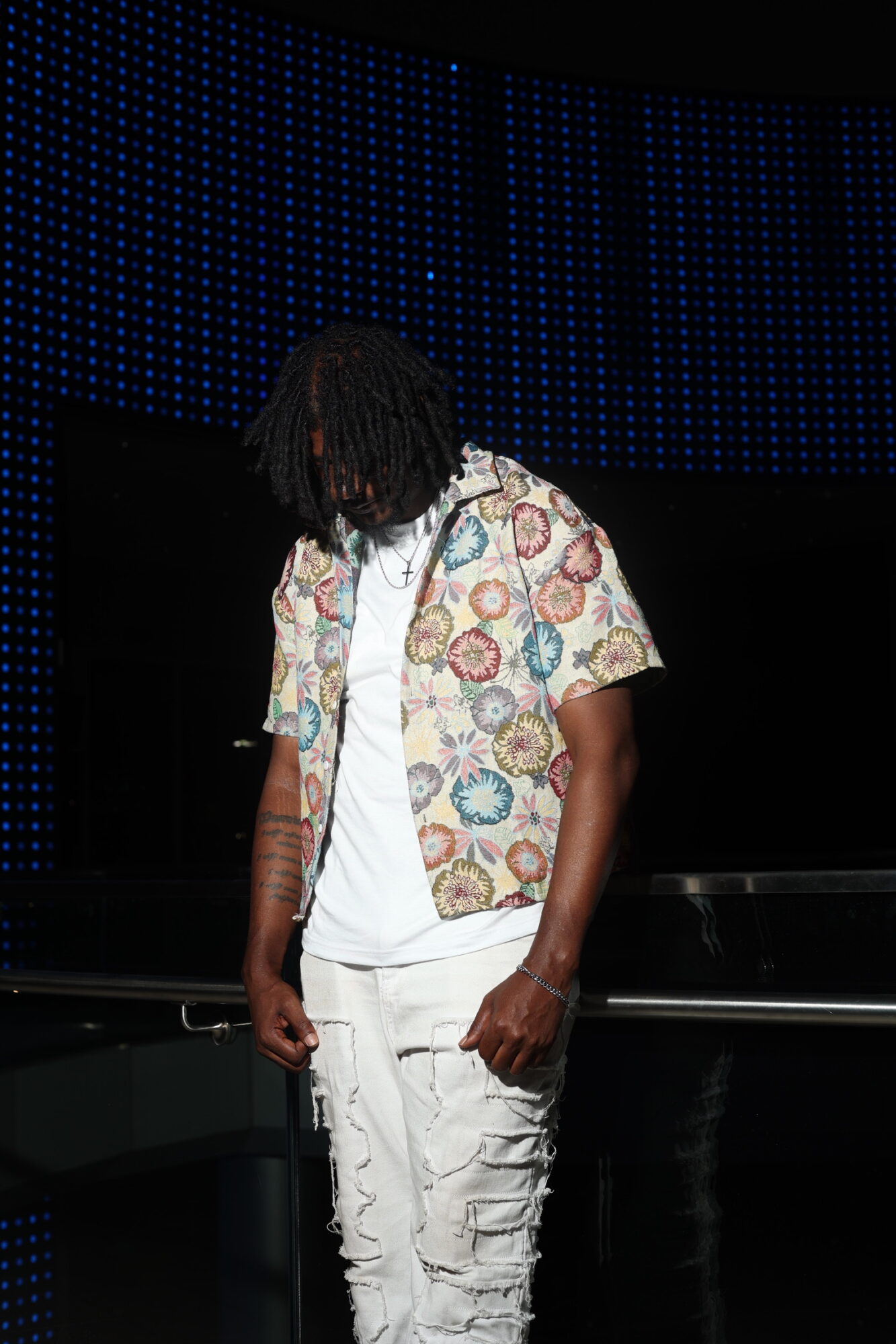 Person with dreadlocks wearing a colorful patterned shirt and white pants, standing on stage with a dark background.