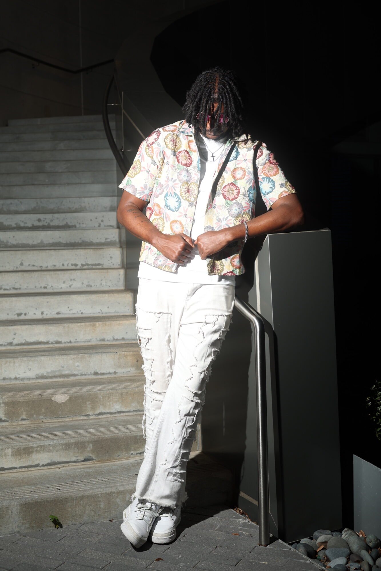Person with dreadlocks wearing a patterned shirt and white pants, standing on stairs, holding a bag strap, looking down.