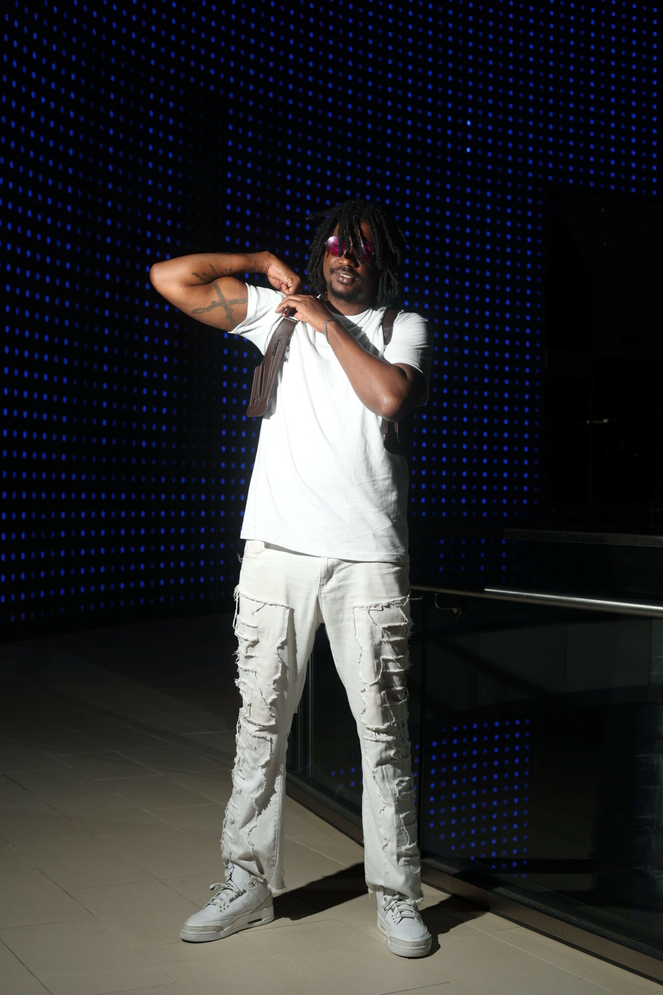 Person standing on a stage with a dark background, holding a microphone, wearing white pants and a white shirt.