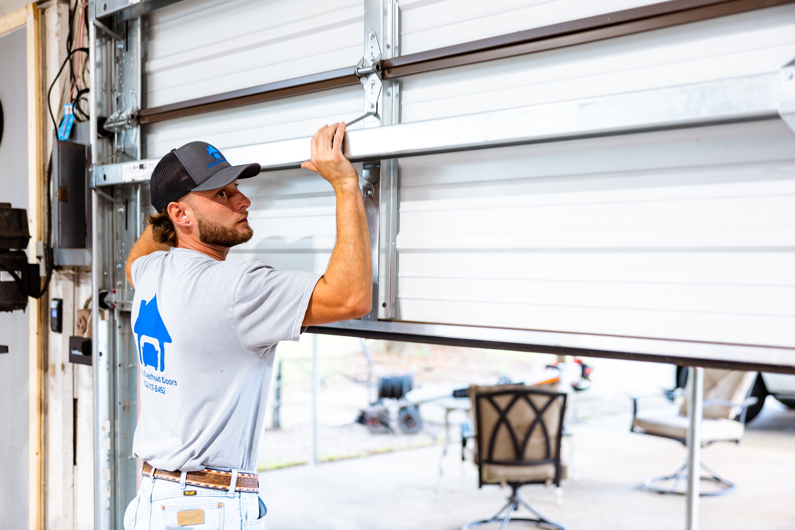 Man in a gray shirt and cap opening a garage door, with a chair and outdoor scene in the background.