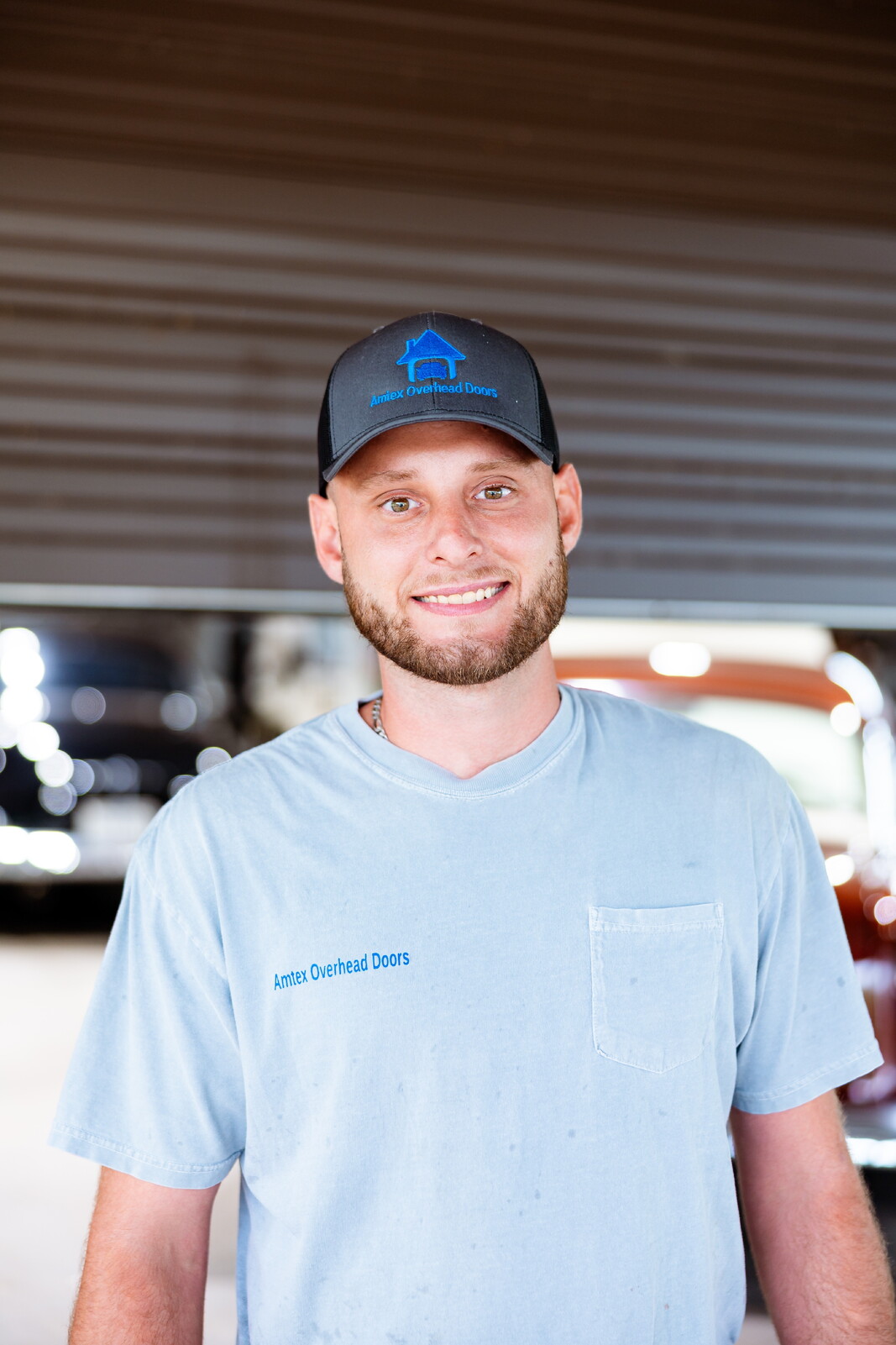 Young man with a beard smiling, wearing a black cap and light blue shirt, standing outdoors under a structure.