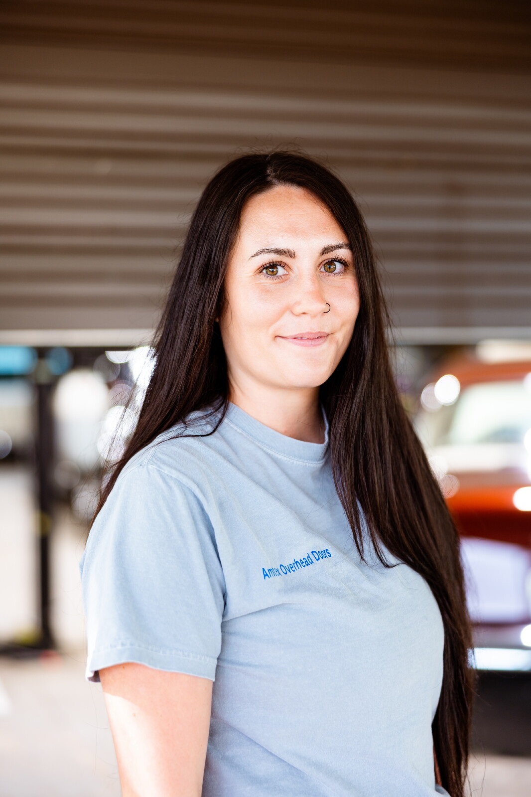 Woman with long dark hair wearing a light blue T-shirt, smiling, in an indoor parking area.