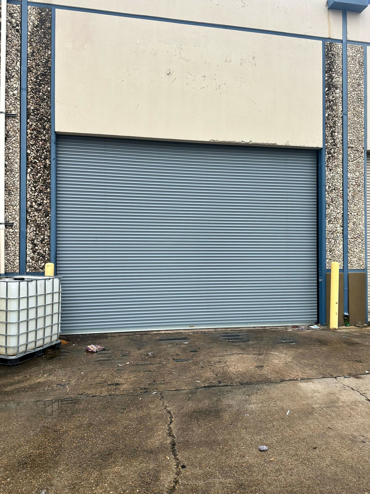Closed blue metal roller shutter door with a white wall on the left and a concrete ground in front.