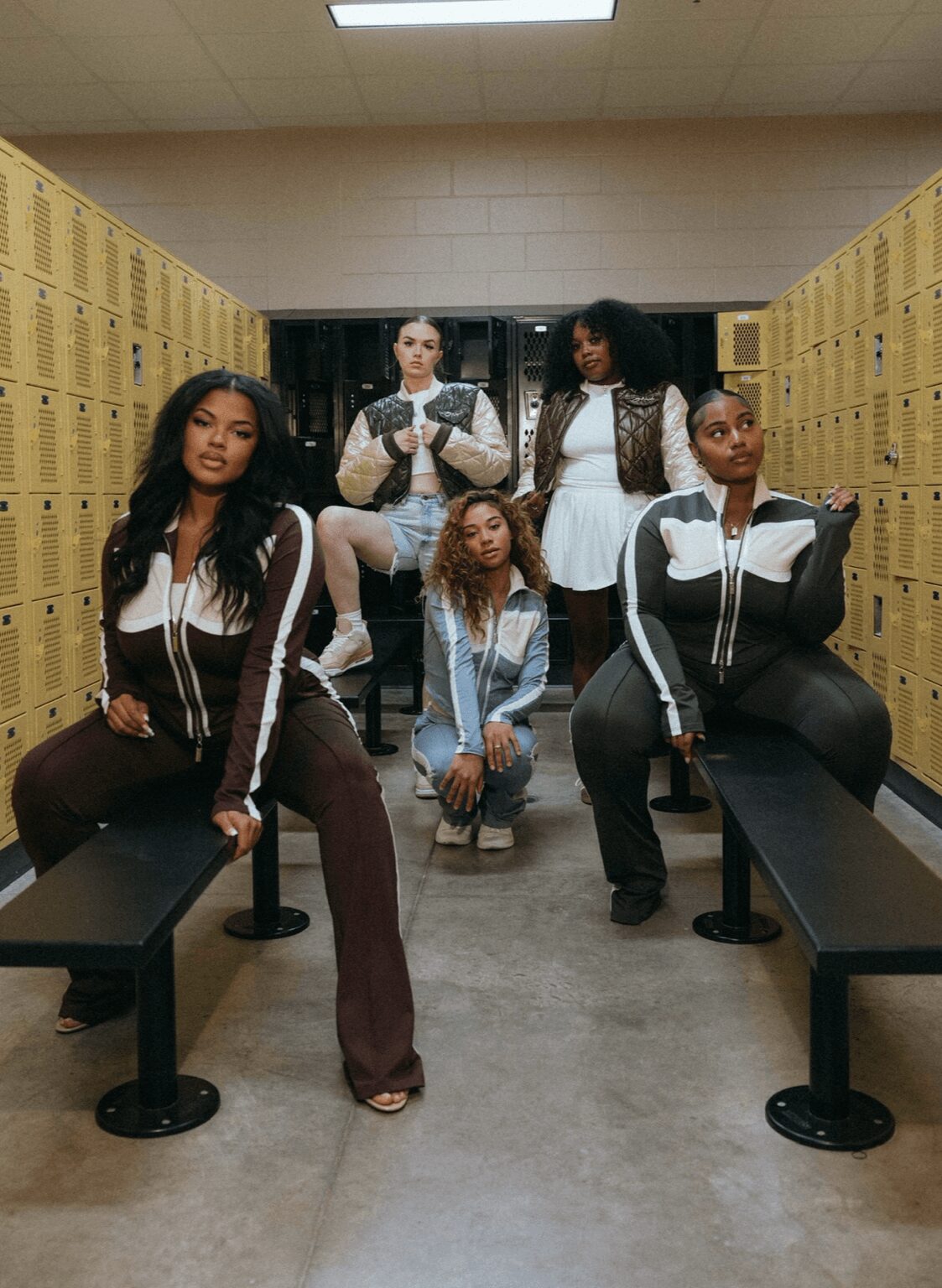 Five women pose in a school hallway with lockers, some sitting on benches and others standing, wearing casual outfits.