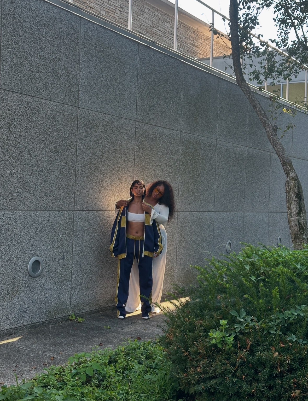 Two people standing on a sidewalk near a gray wall, with a tree and greenery nearby.