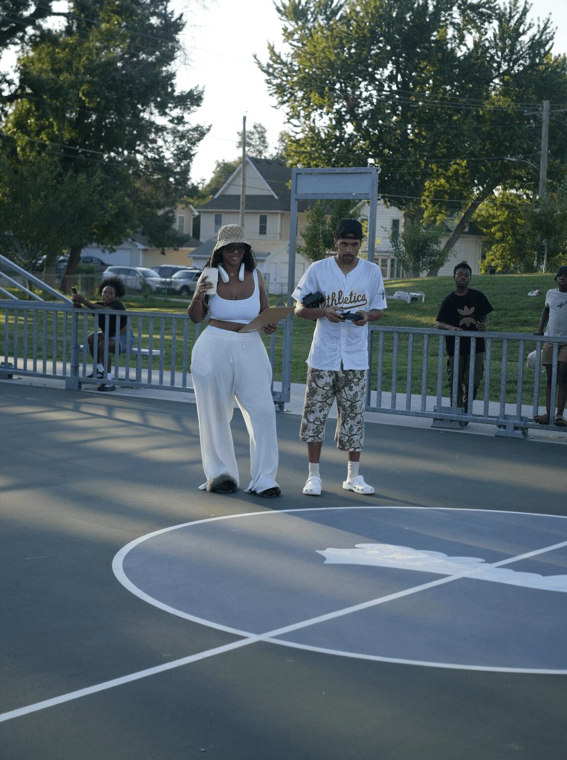 Two people standing on a basketball court, with a group of people and trees in the background.