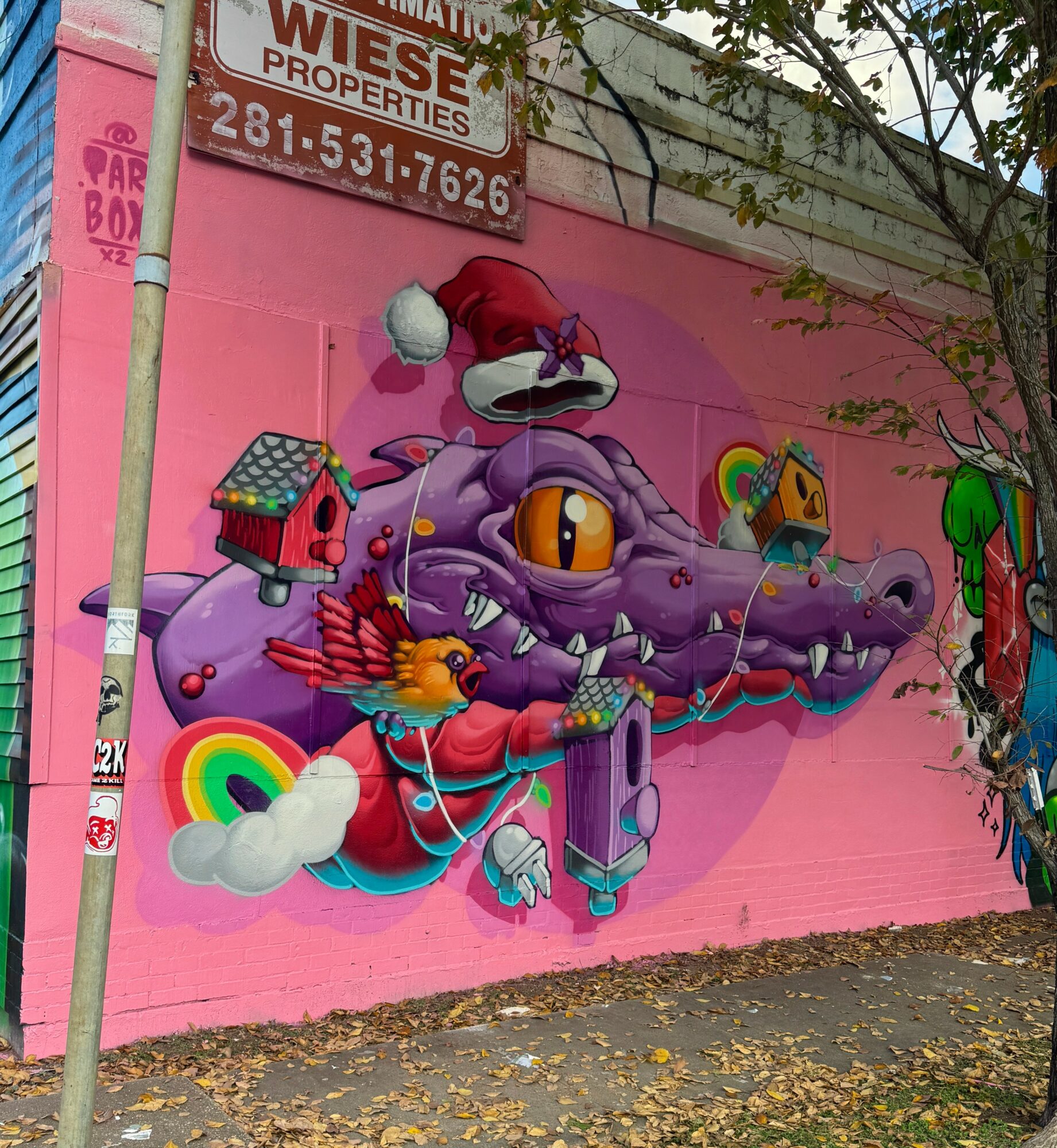 Colorful mural of a purple crocodile with a rainbow, house, and bird on pink wall, with tree and sign nearby.