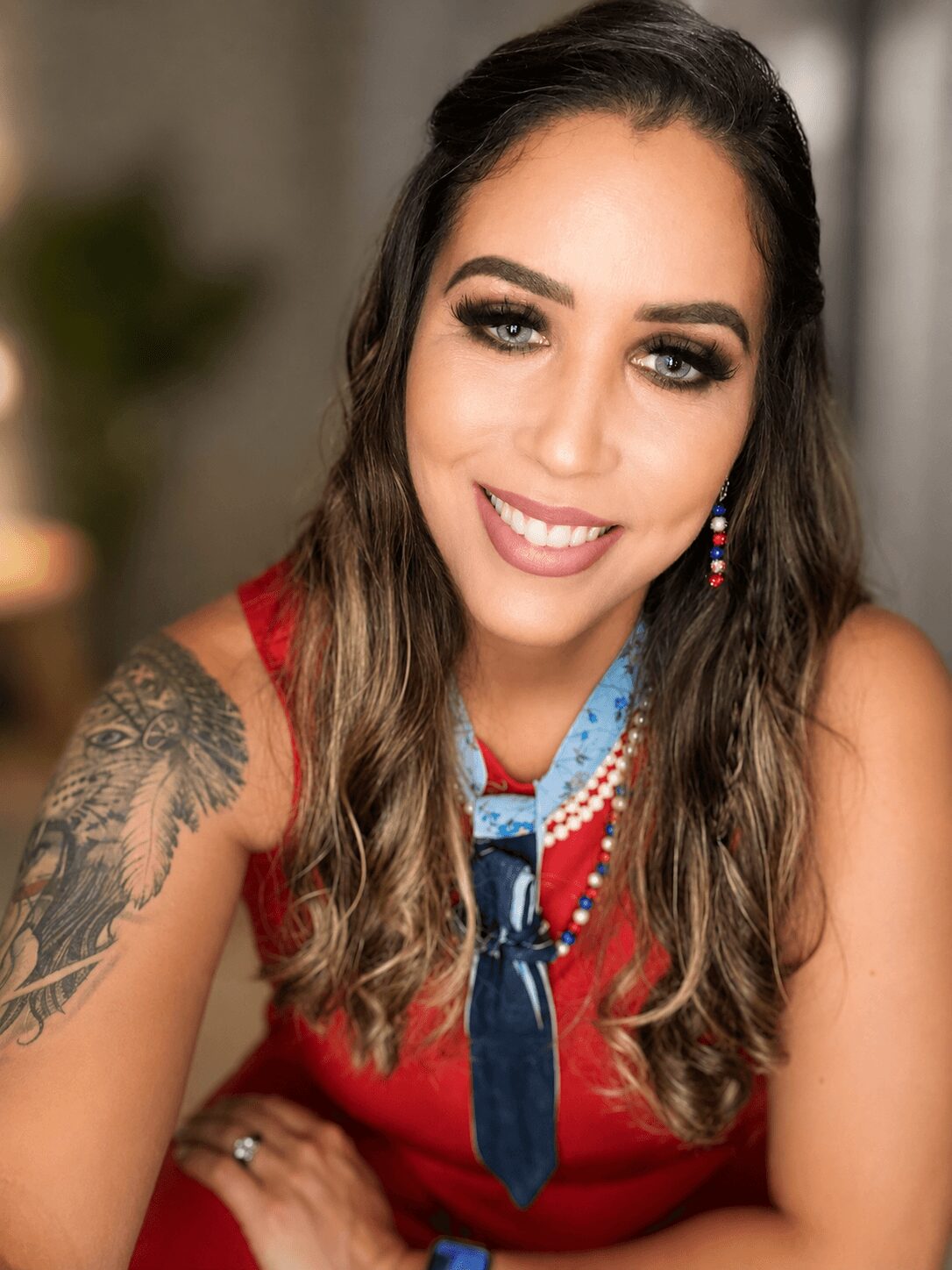 Smiling woman with long wavy hair, makeup, and colorful earrings, wearing a red top with a blue scarf, in an indoor setting.