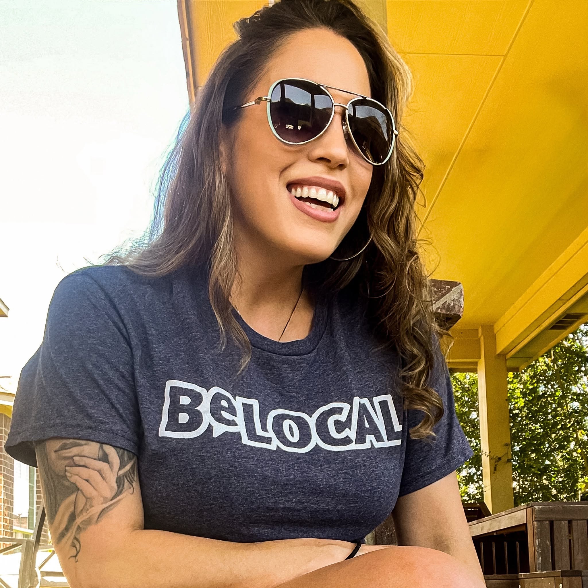 Woman with sunglasses smiling, wearing a dark t-shirt, outdoors with yellow structure and trees in background.