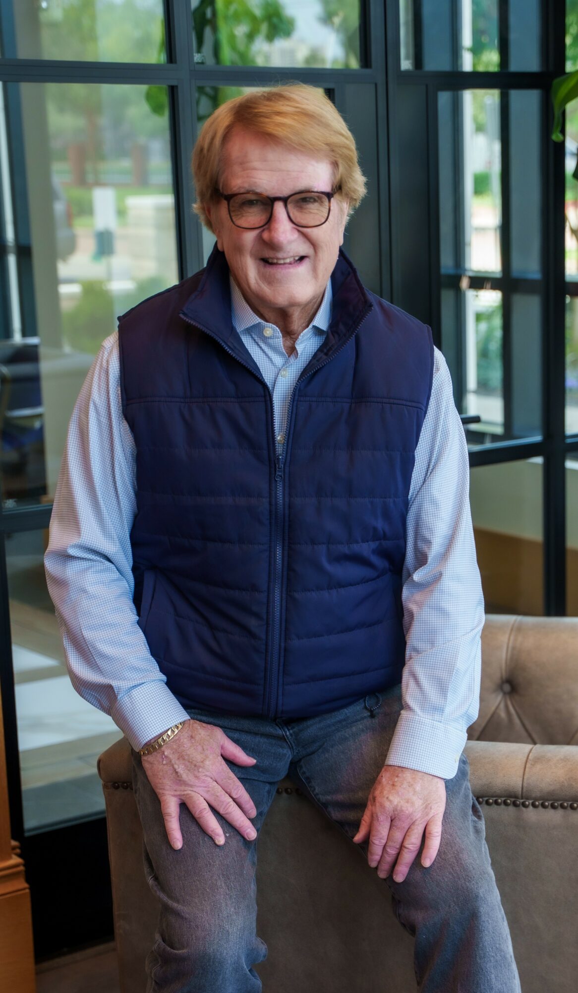 Older man with glasses, smiling, wearing a navy vest over a light blue shirt, standing indoors near a window.