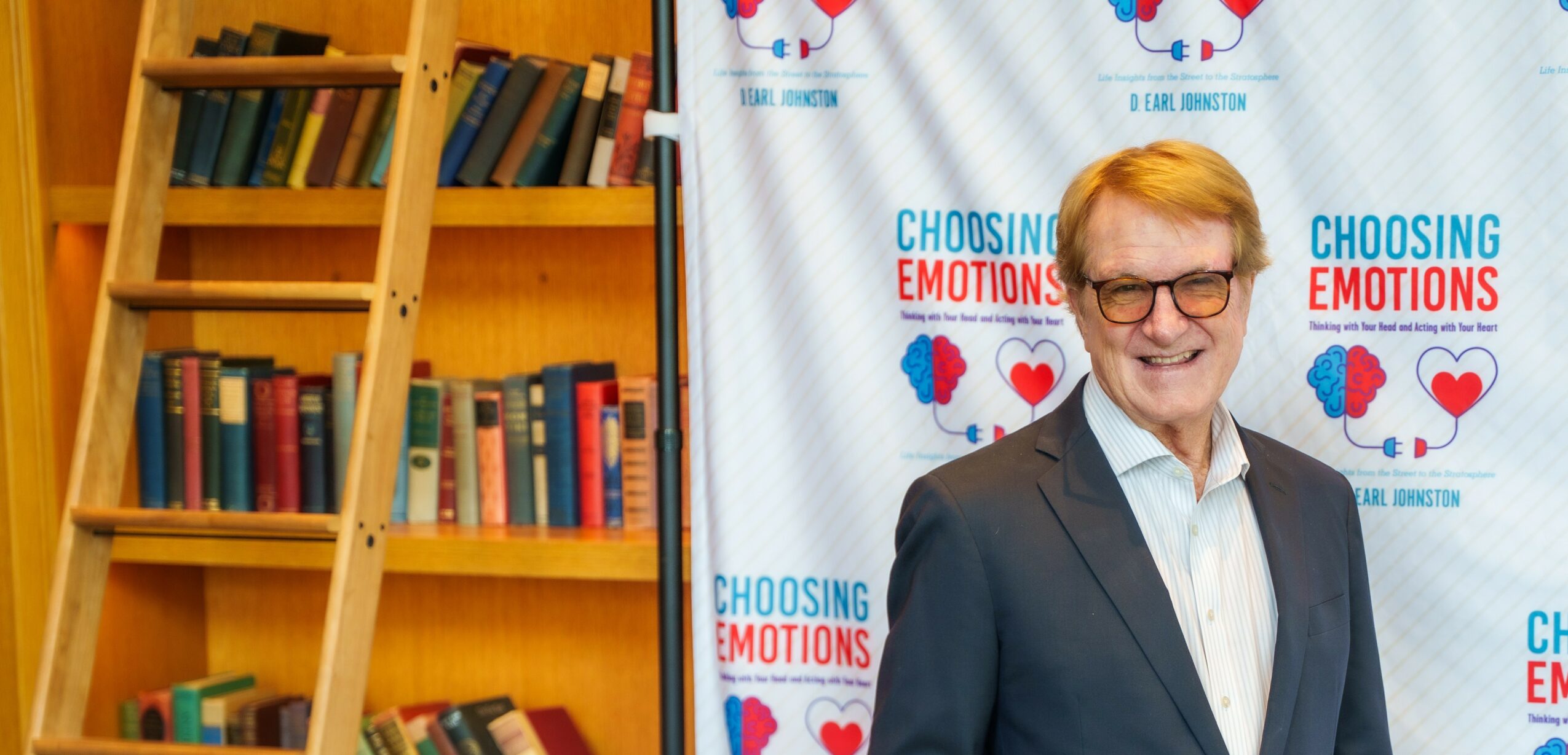 Smiling man in glasses and suit standing in front of a backdrop with 'Choosing Emotions' text and heart graphics.