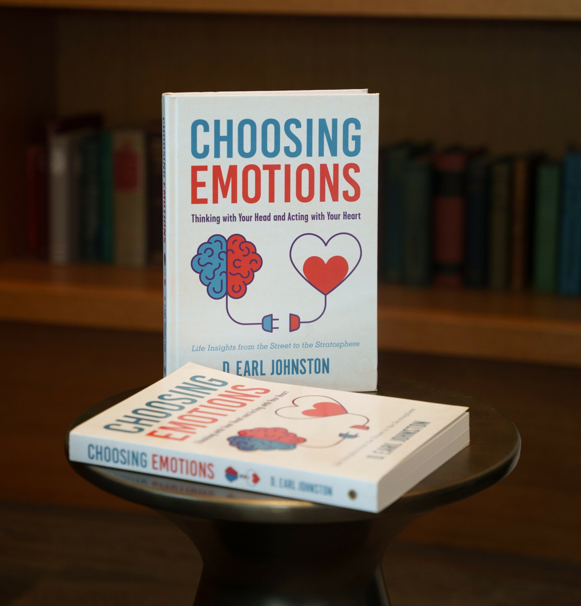Two copies of a book titled 'Choosing Emotions' on a black round table, with a bookshelf in the background.