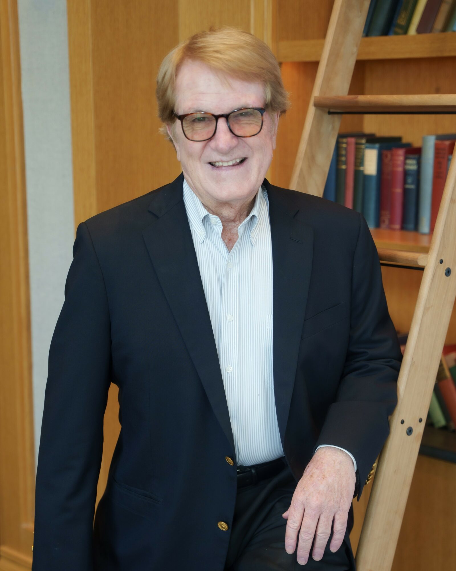 Older man with glasses, smiling, wearing a dark blazer and white shirt, standing indoors near a wooden bookshelf and ladder.