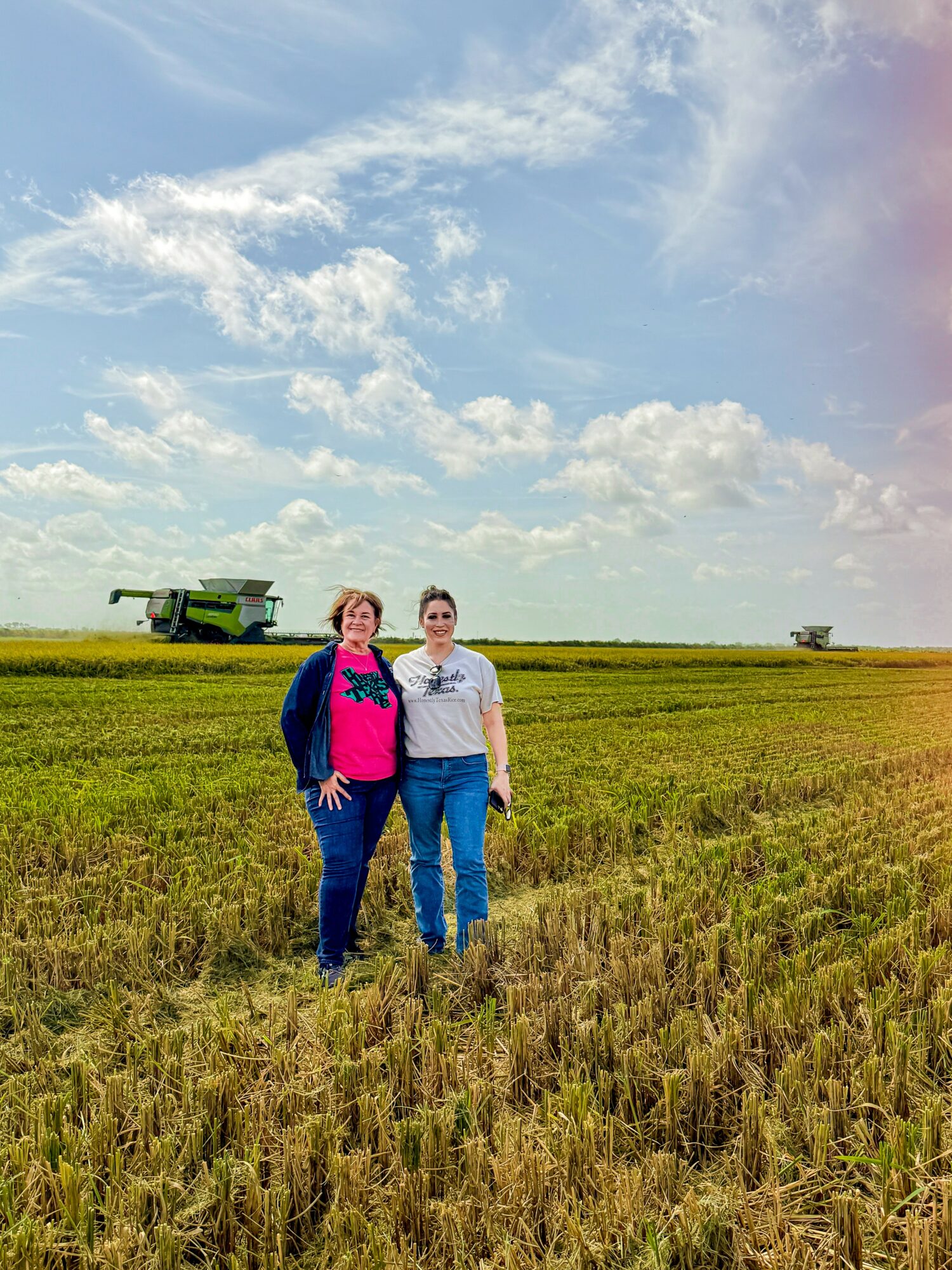 Two women standing in a field under a partly cloudy sky, with a combine harvester in the background.