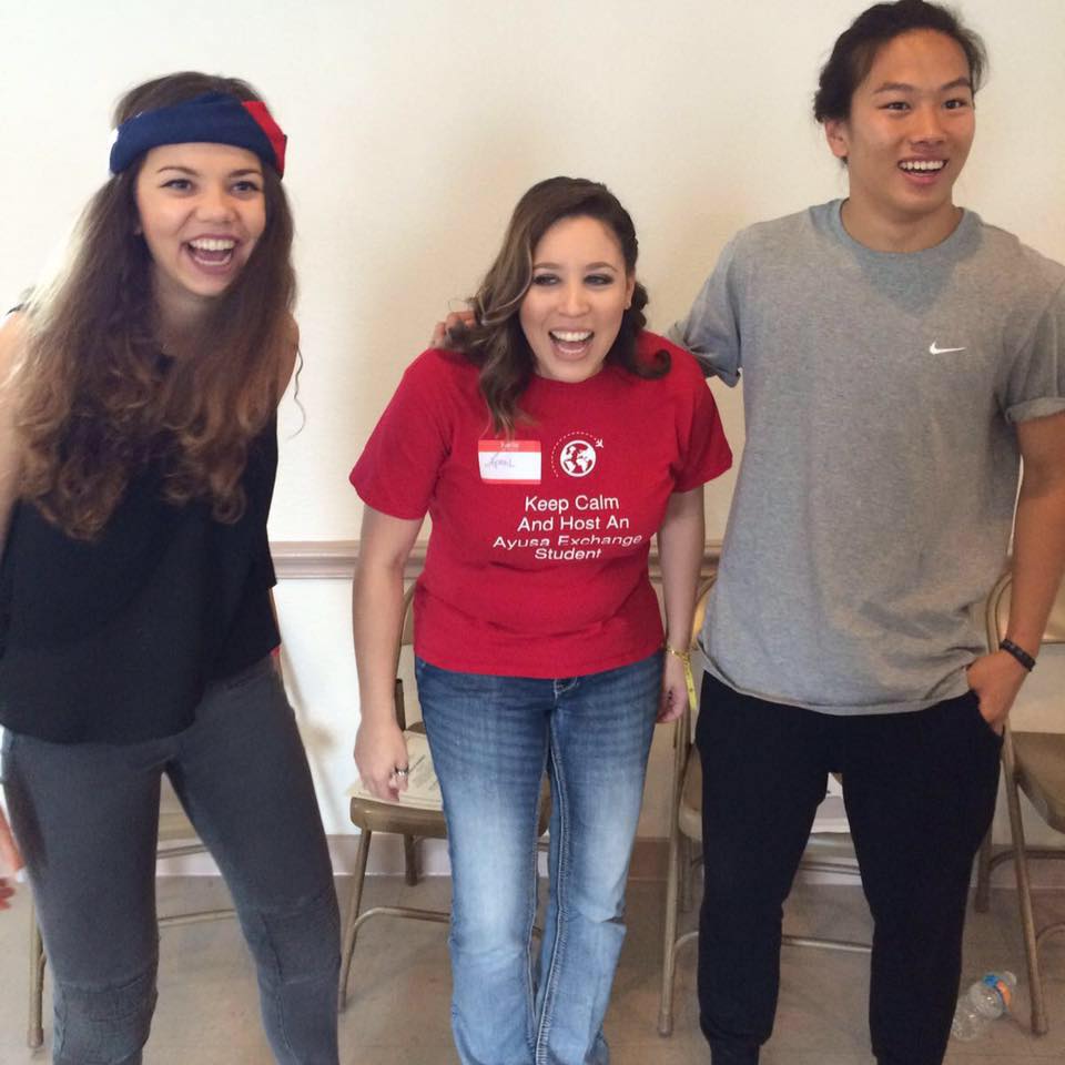 Three people standing together, smiling, in a room with chairs, one wearing a red shirt, one in gray, one in black, casual setting.